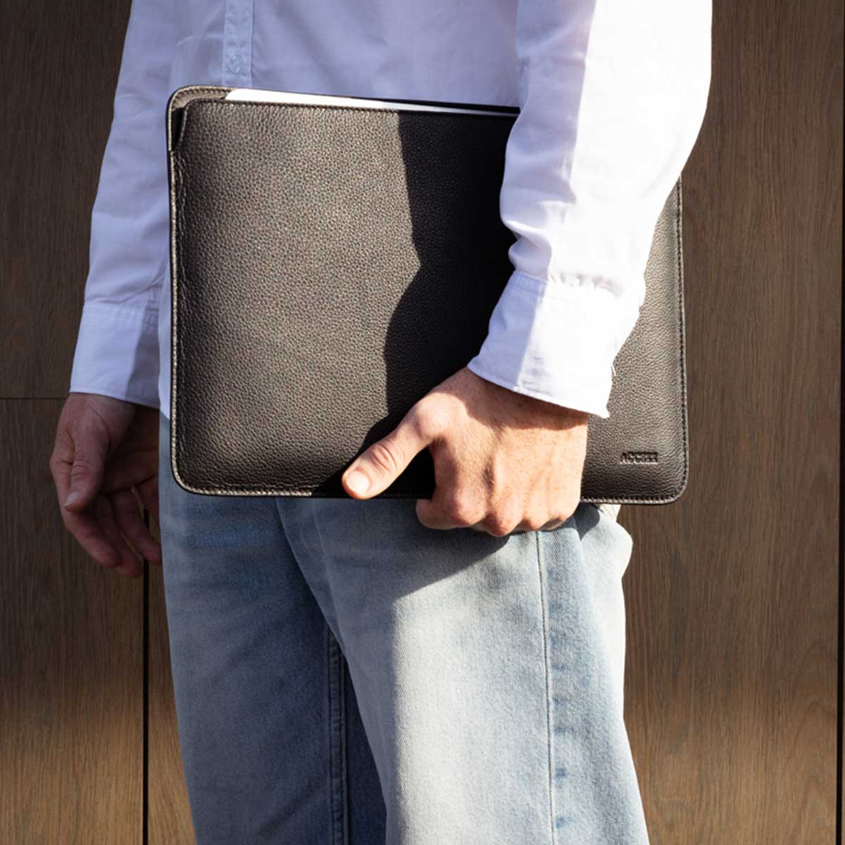 A person holding a black leather laptop sleeve, against a wooden background. The sleeve is closed.