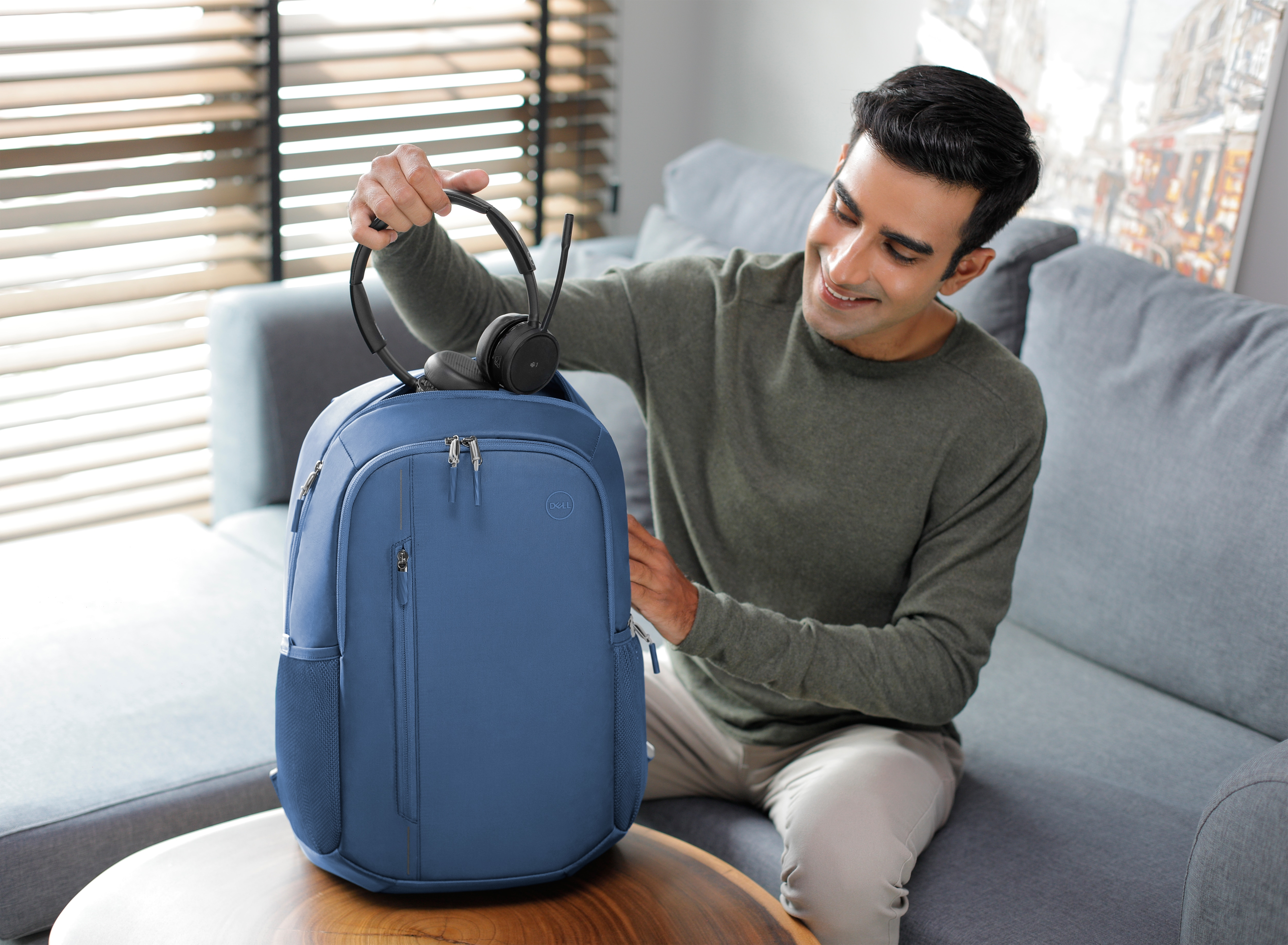 Mann lächelt, hält ein schwarzes Headset und öffnet einen blauen Rucksack.