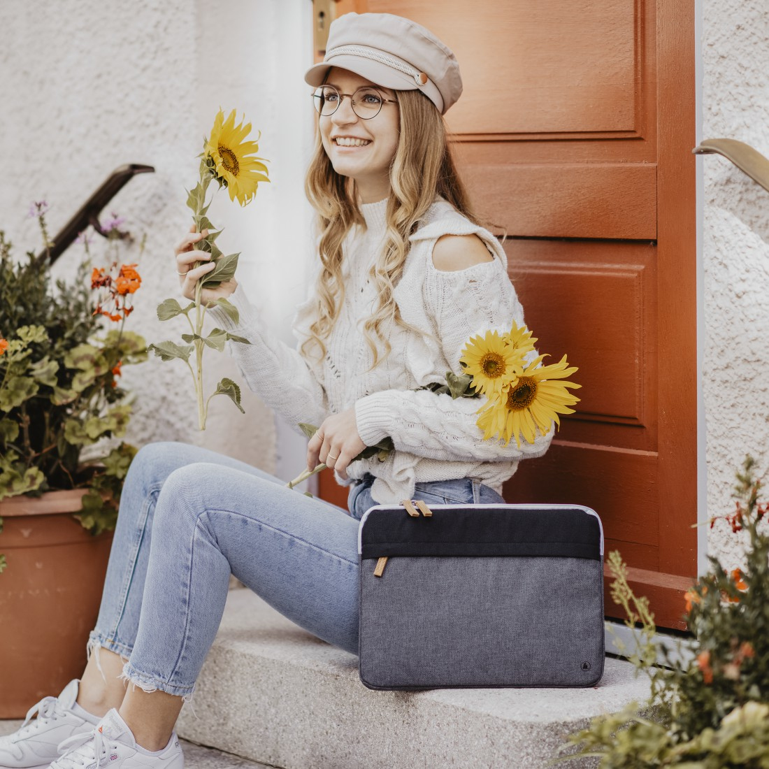 Kobieta z słonecznikami, siedząca na schodach, z etui na laptopa. Widoczne drzwi i rośliny.