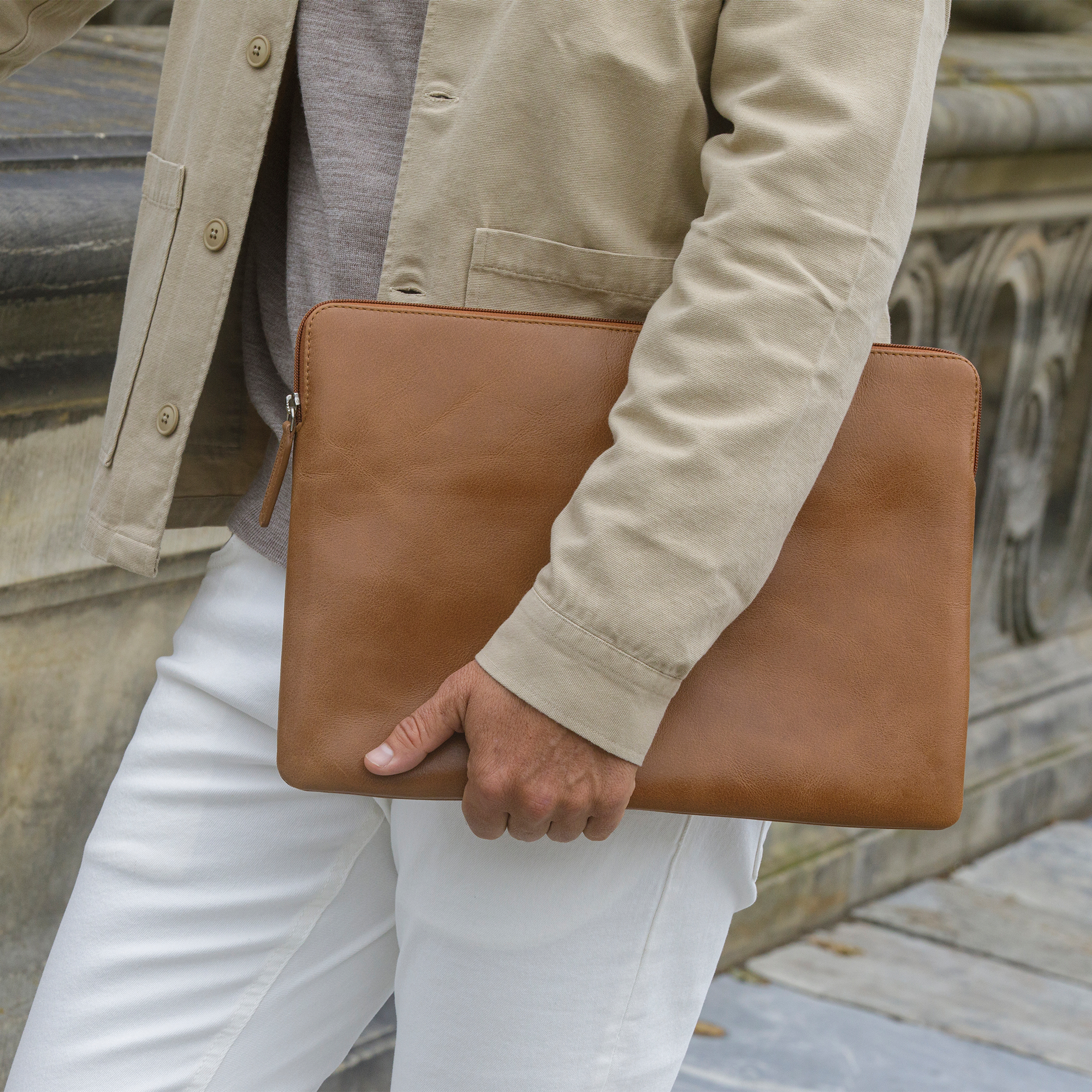 Persona che tiene una custodia per laptop in pelle marrone. Giacca beige, pantaloni bianchi visibili.
