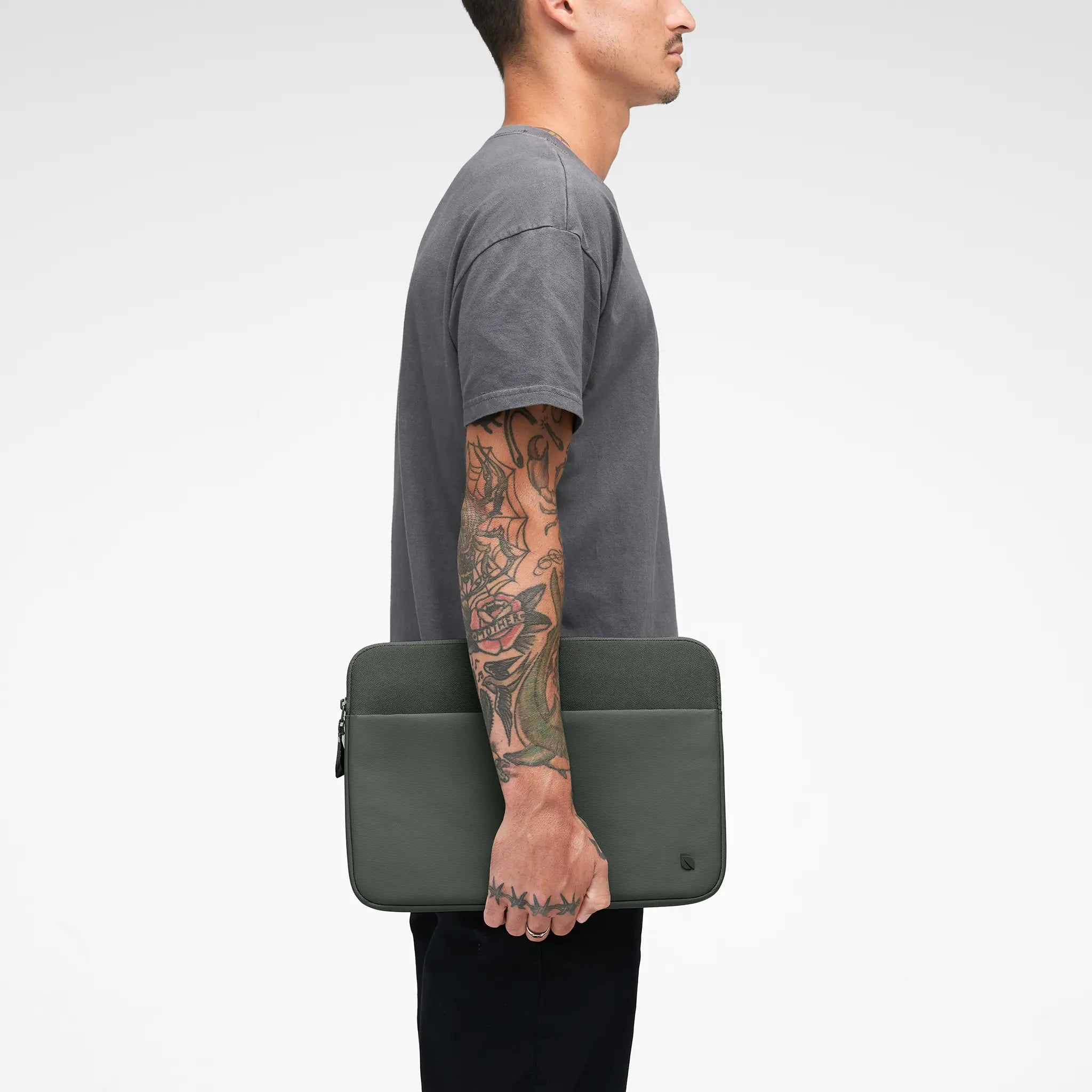 Uomo che tiene una custodia grigia per laptop, vista laterale, con tatuaggi sul braccio.