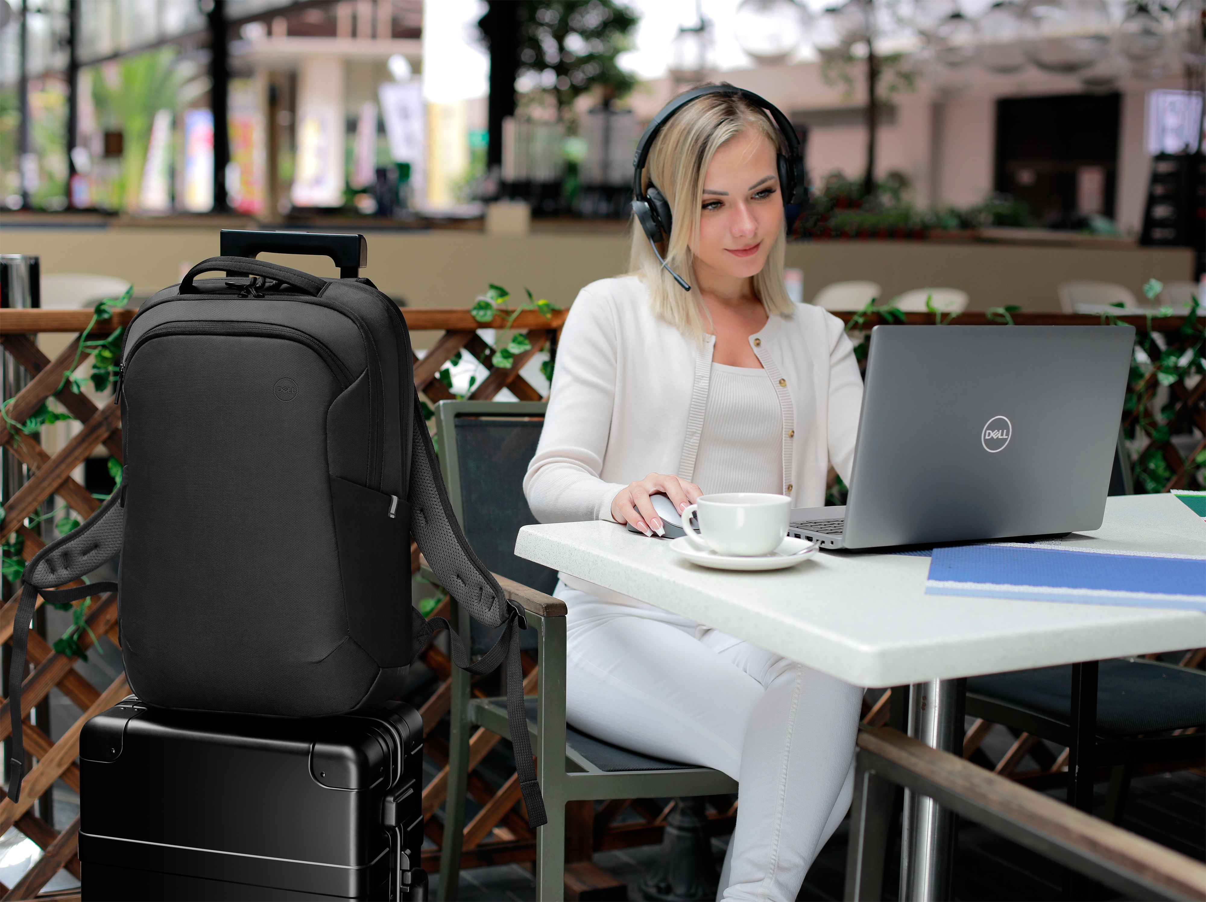 Frau benutzt Laptop, trägt Headset. Schwarzer Rucksack und Koffer links.