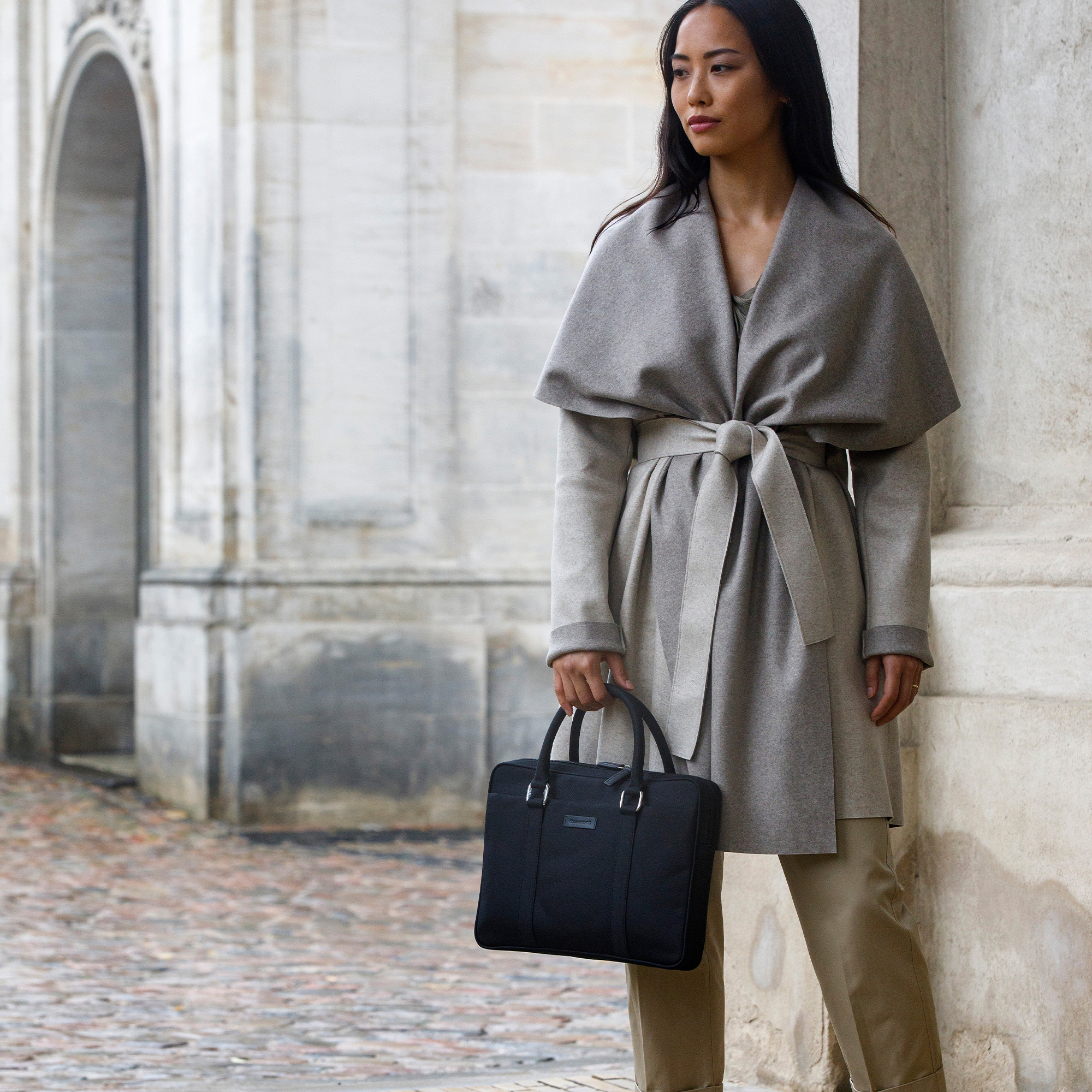 Donna con cappotto grigio che tiene una ventiquattrore nera, vicino a un edificio.