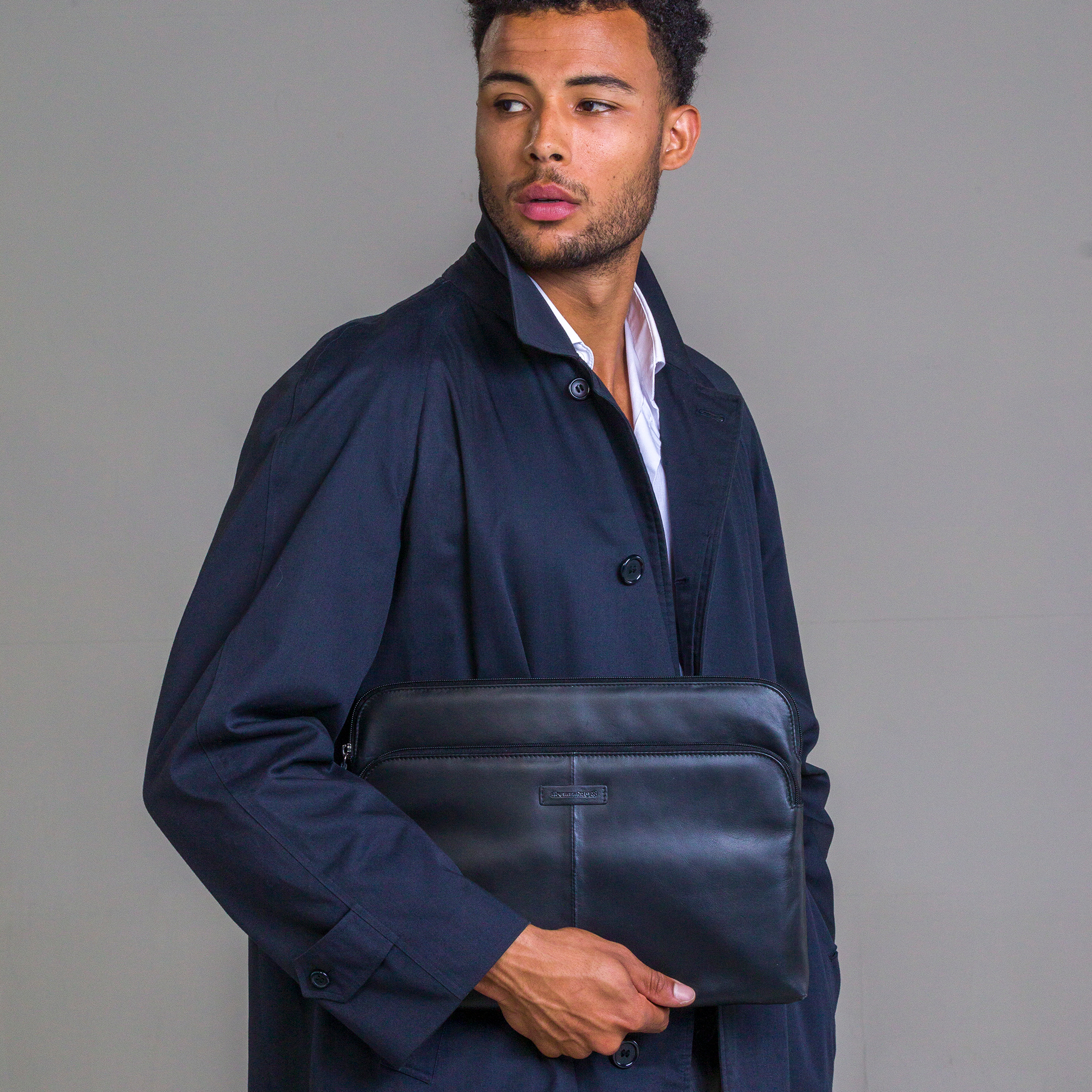 Un homme tenant une housse d'ordinateur portable en cuir noir, portant un manteau sombre et une chemise blanche, fond gris.