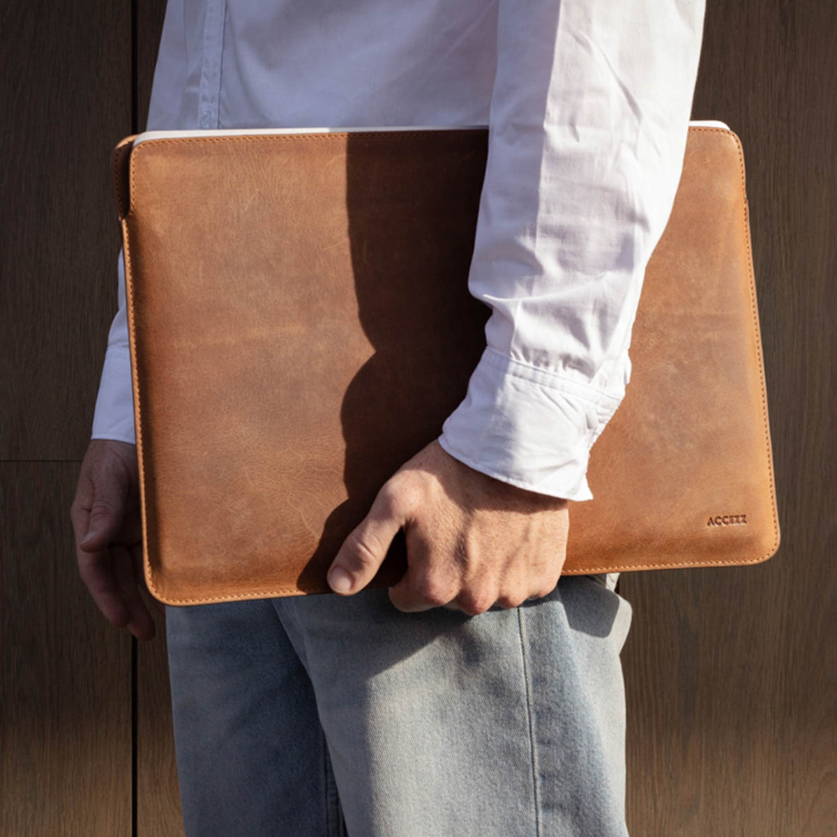 Ein Mann hält eine braune Laptop-Hülle aus Leder. Weißes Hemd und Jeans.