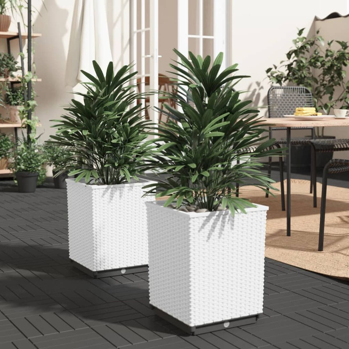 Zwei weiße quadratische Pflanzgefäße mit grünen Pflanzen auf einer Terrasse. Tisch und Stühle im Hintergrund.