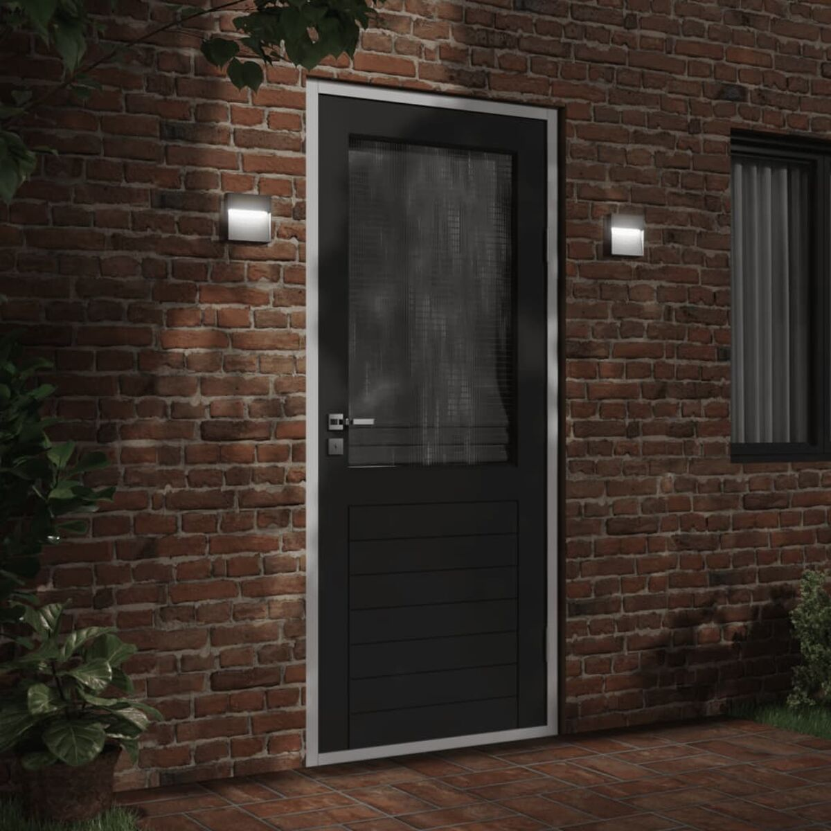 Schwarze Tür mit Fenster, Backsteinmauer, zwei Wandlampen und Pflanzen am Boden.