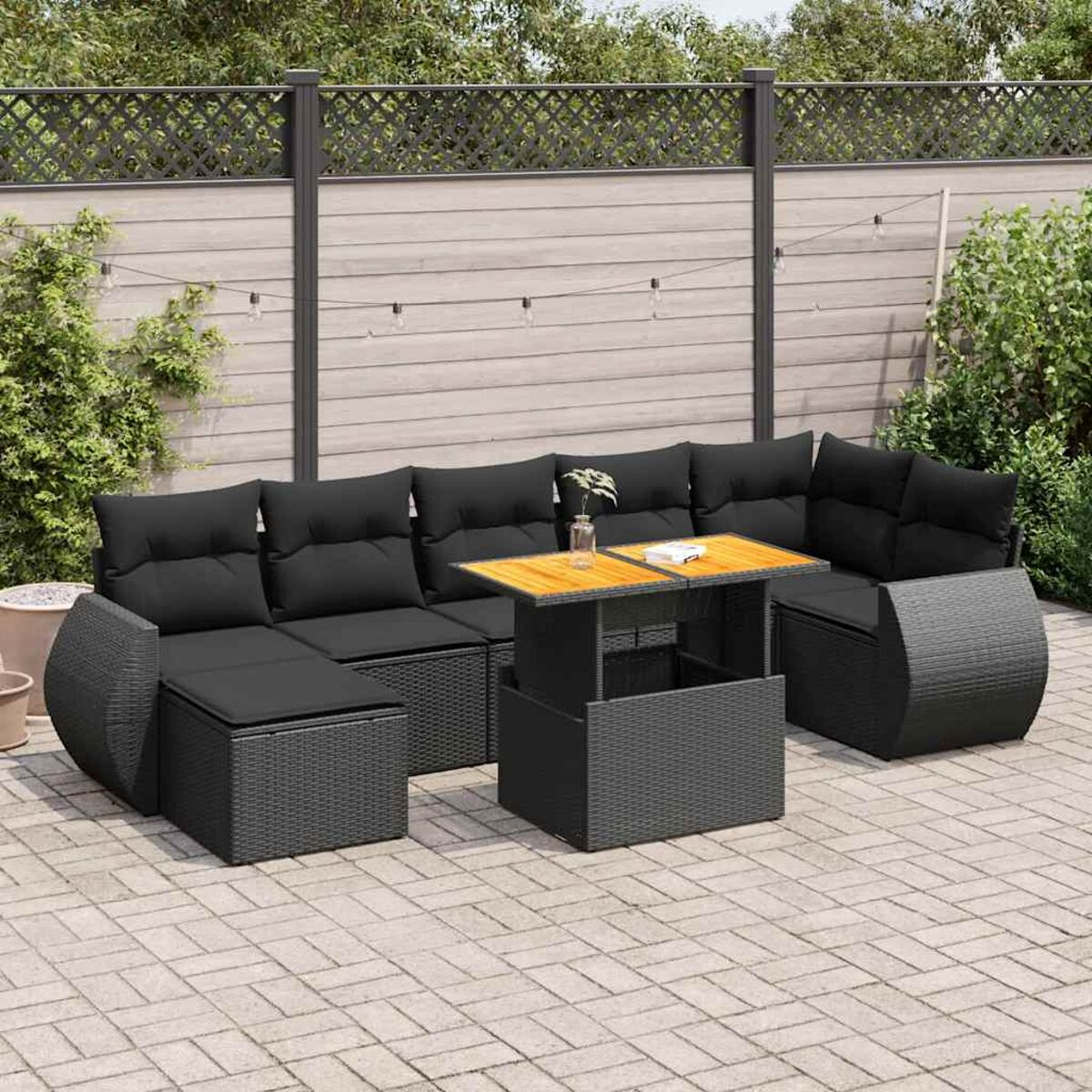 Set di mobili da esterno con cuscini neri e tavolo sollevabile. Patio pavimentato e vegetazione sullo sfondo.
