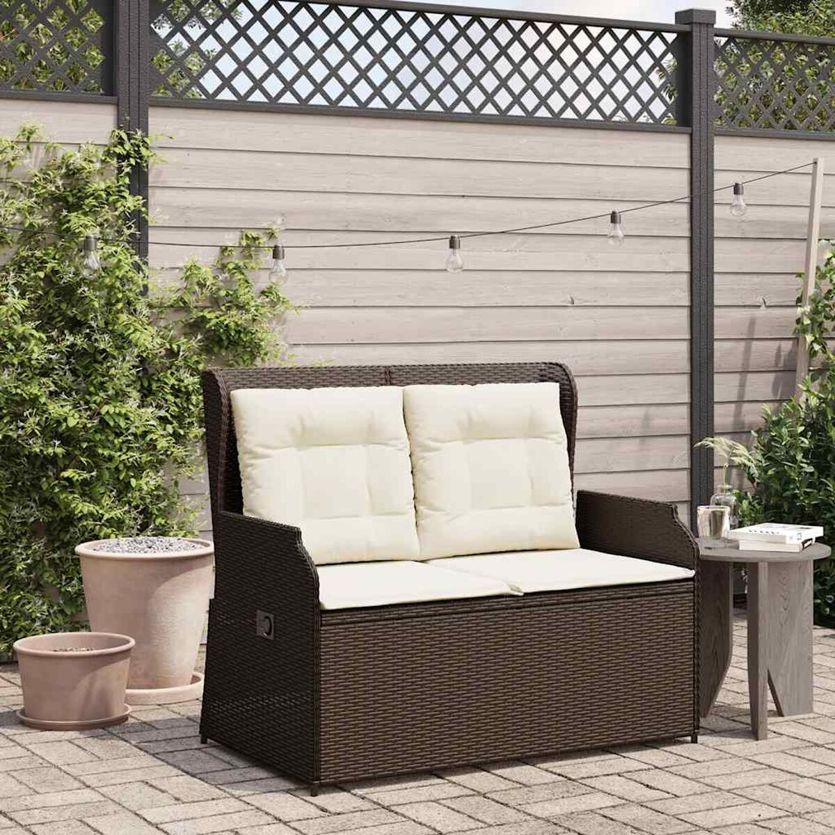 Una panchina in vimini marrone con cuscini color crema in un giardino con una recinzione grigia.
