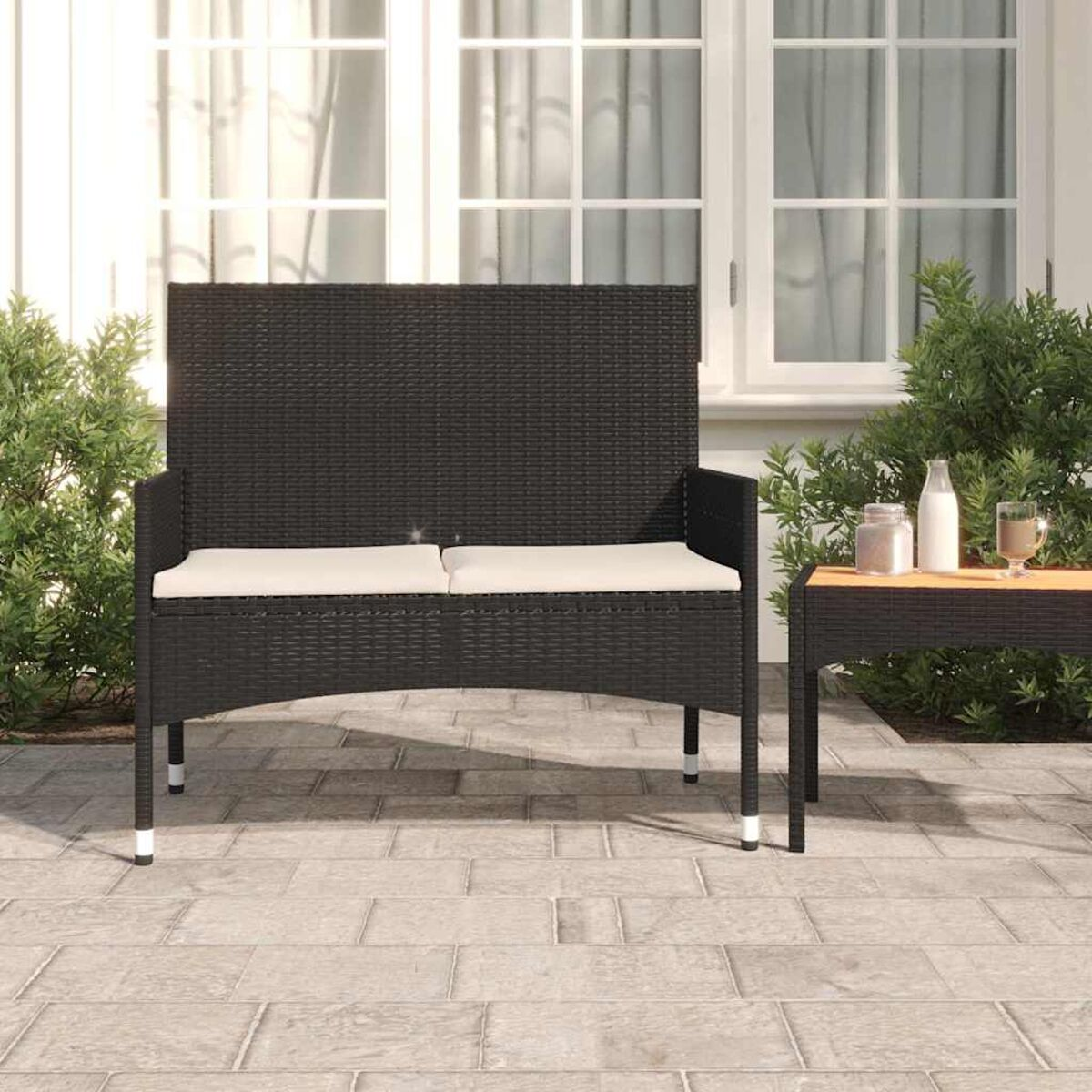 Schwarze Outdoor-Bank mit weißen Kissen, Tisch mit Getränken und Büschen im Hintergrund.