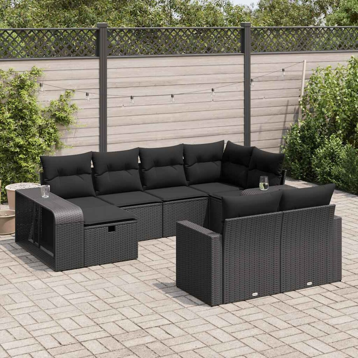 Set patio in vimini con cuscini neri, tavolo e poltrona su patio in mattoni, staccionata in legno sullo sfondo.