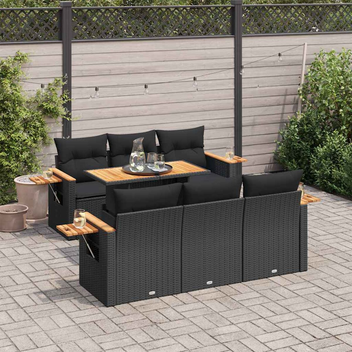 Set di sedute da esterno in rattan nero con tavolo in legno e cuscini neri, patio in mattoni e luci.