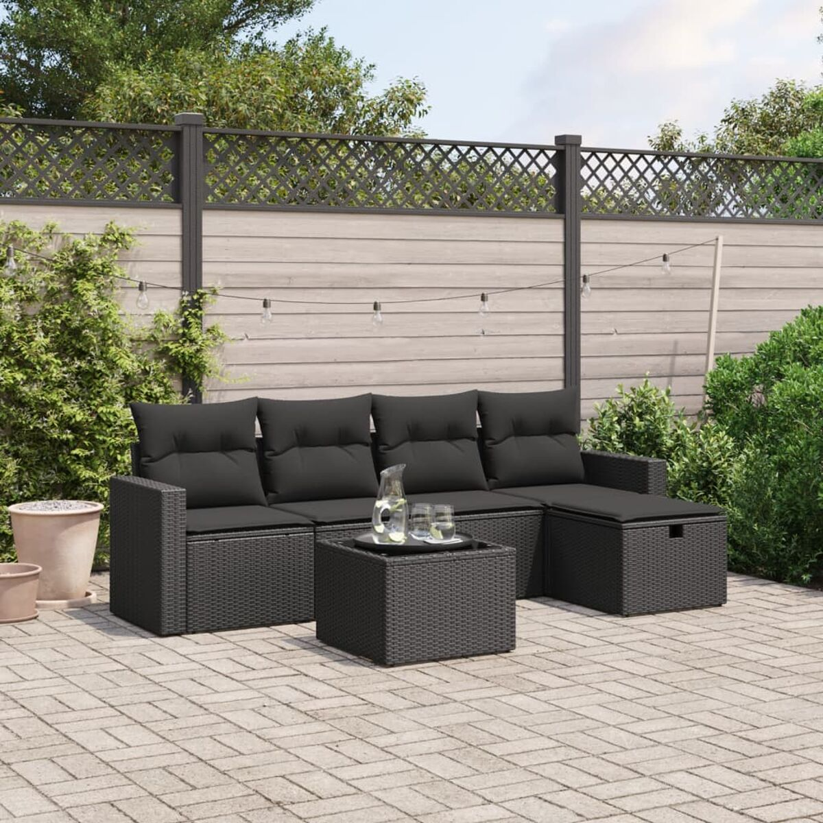 Schwarzes Korbmöbel-Set für die Terrasse, Sofa, Ottomane und Couchtisch auf einer Backsteinterrasse mit Grün.