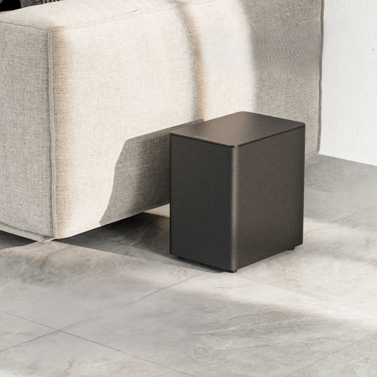 Czarny głośnik obok jasnobeżowej sofy. Głośnik stoi na szarej podłodze z płytek.