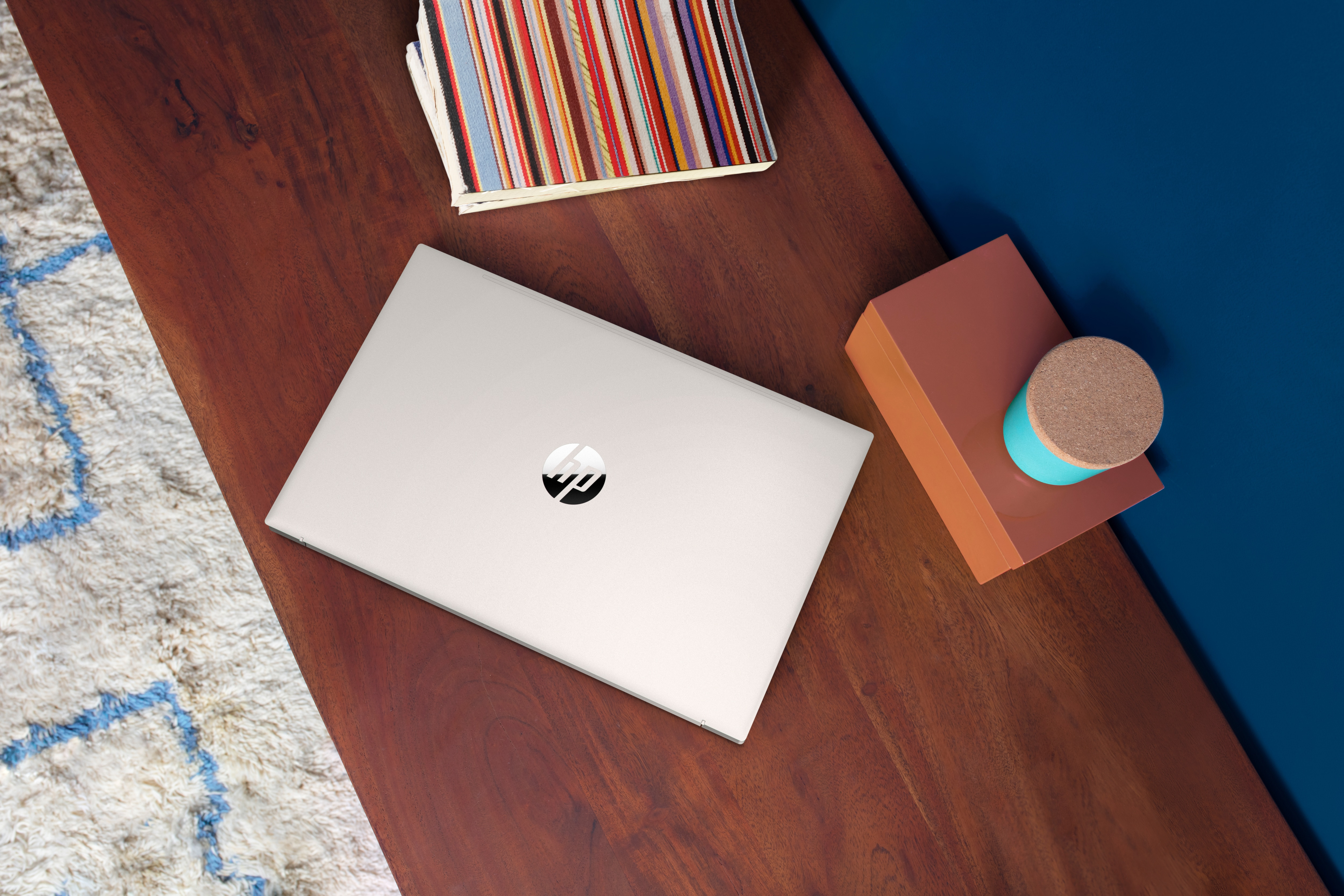 Laptop na drewnianym stole z wzorzystym dywanem, stosem książek i brązowym pudełkiem z walcem.