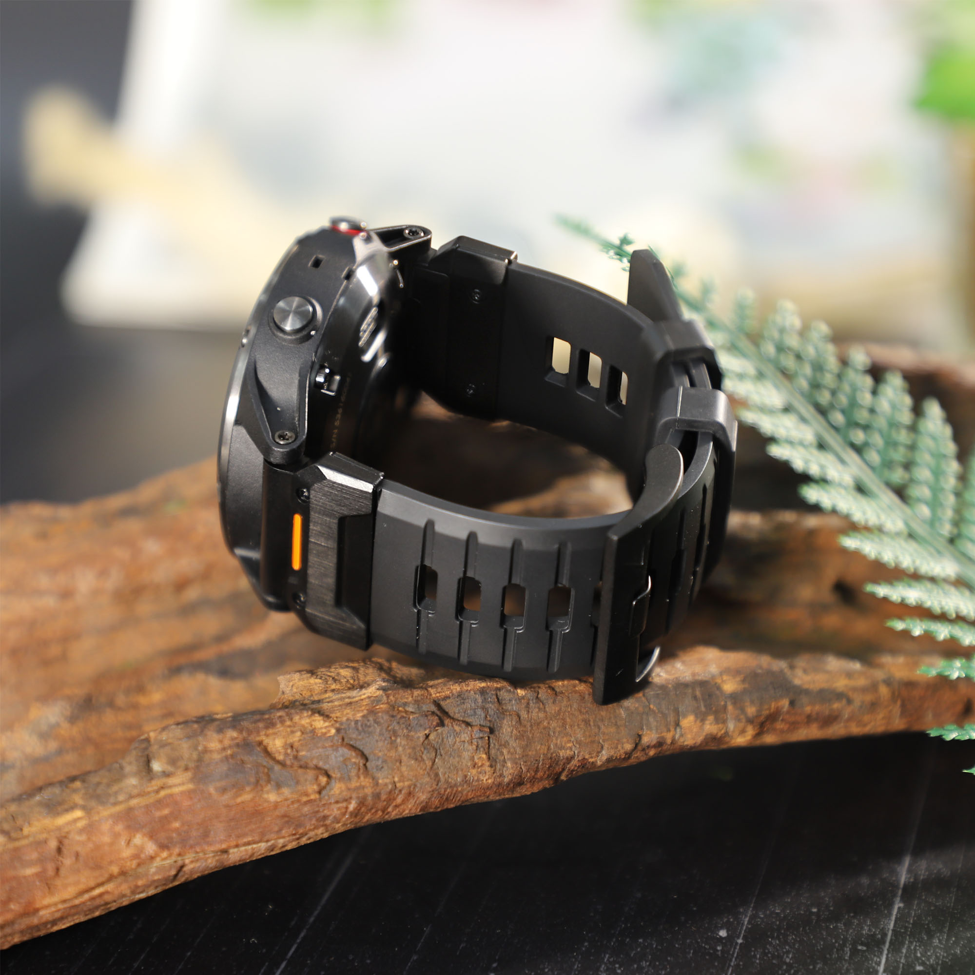 Schwarze Smartwatch auf Holz mit einer grünen Pflanze im Hintergrund. Das Armband ist ebenfalls schwarz.