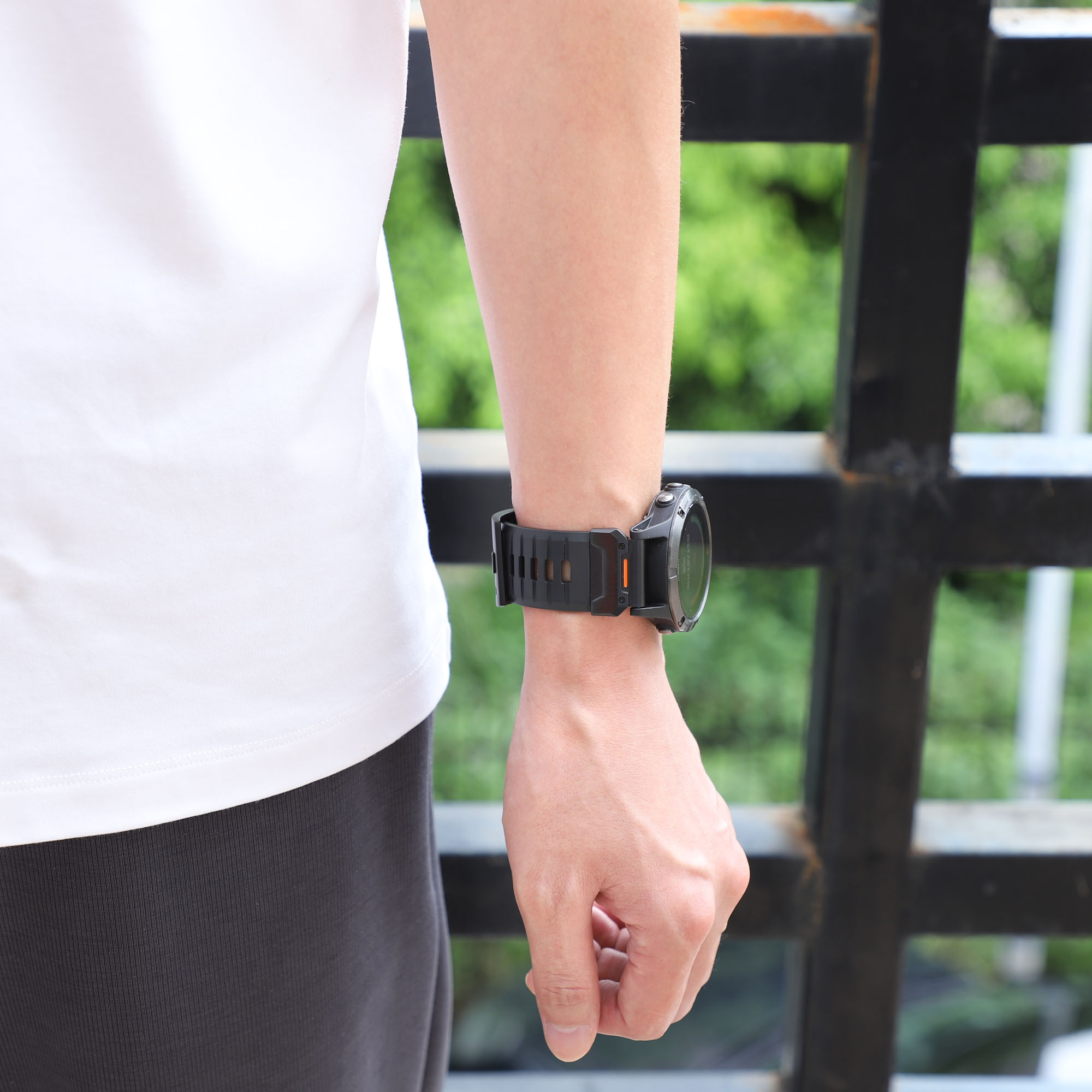 Eine Person, die eine schwarz-orangefarbene Smartwatch am Handgelenk trägt, auf einer Brücke mit Grün.