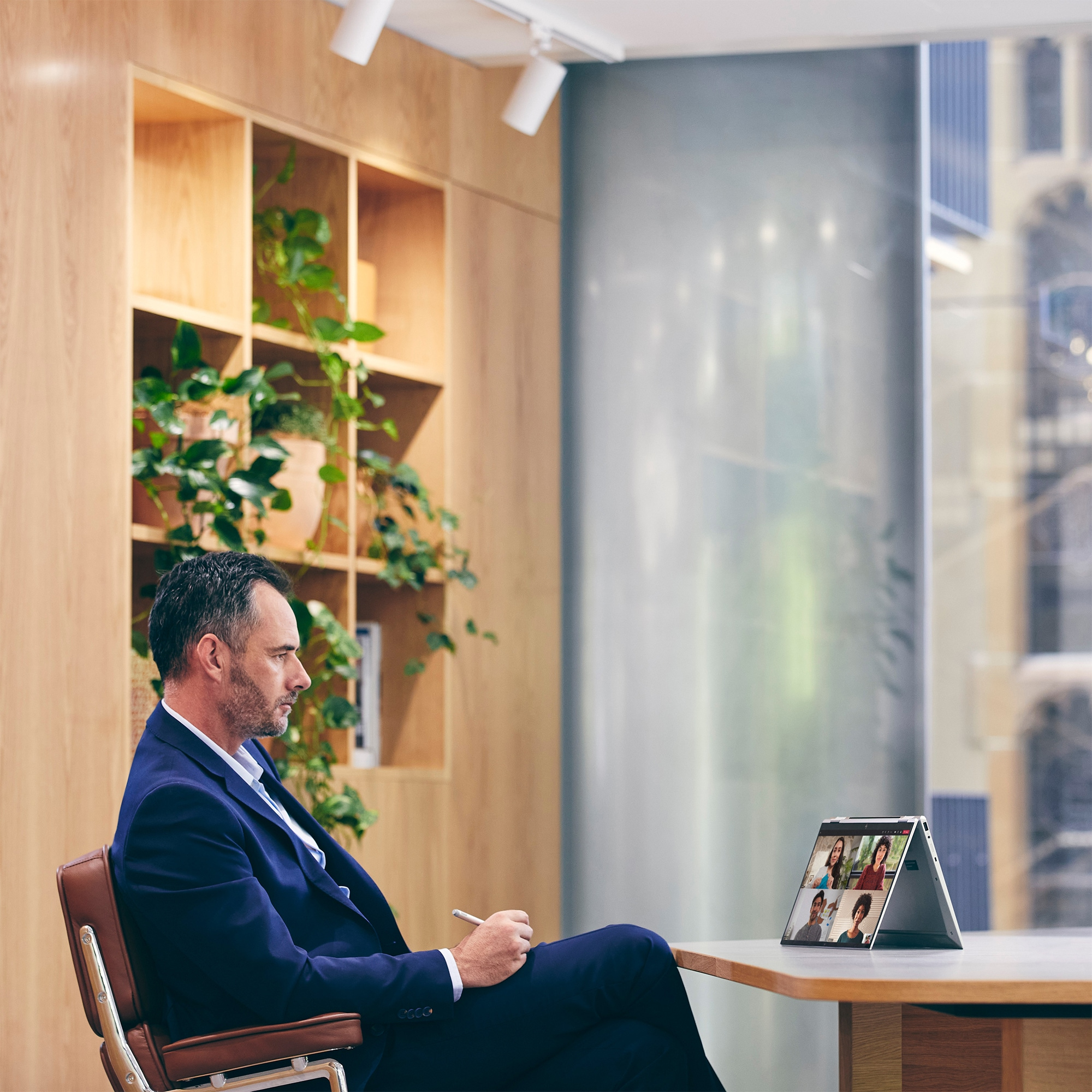 A man in a suit is on a video call on a laptop. Plants and a window are in the background.