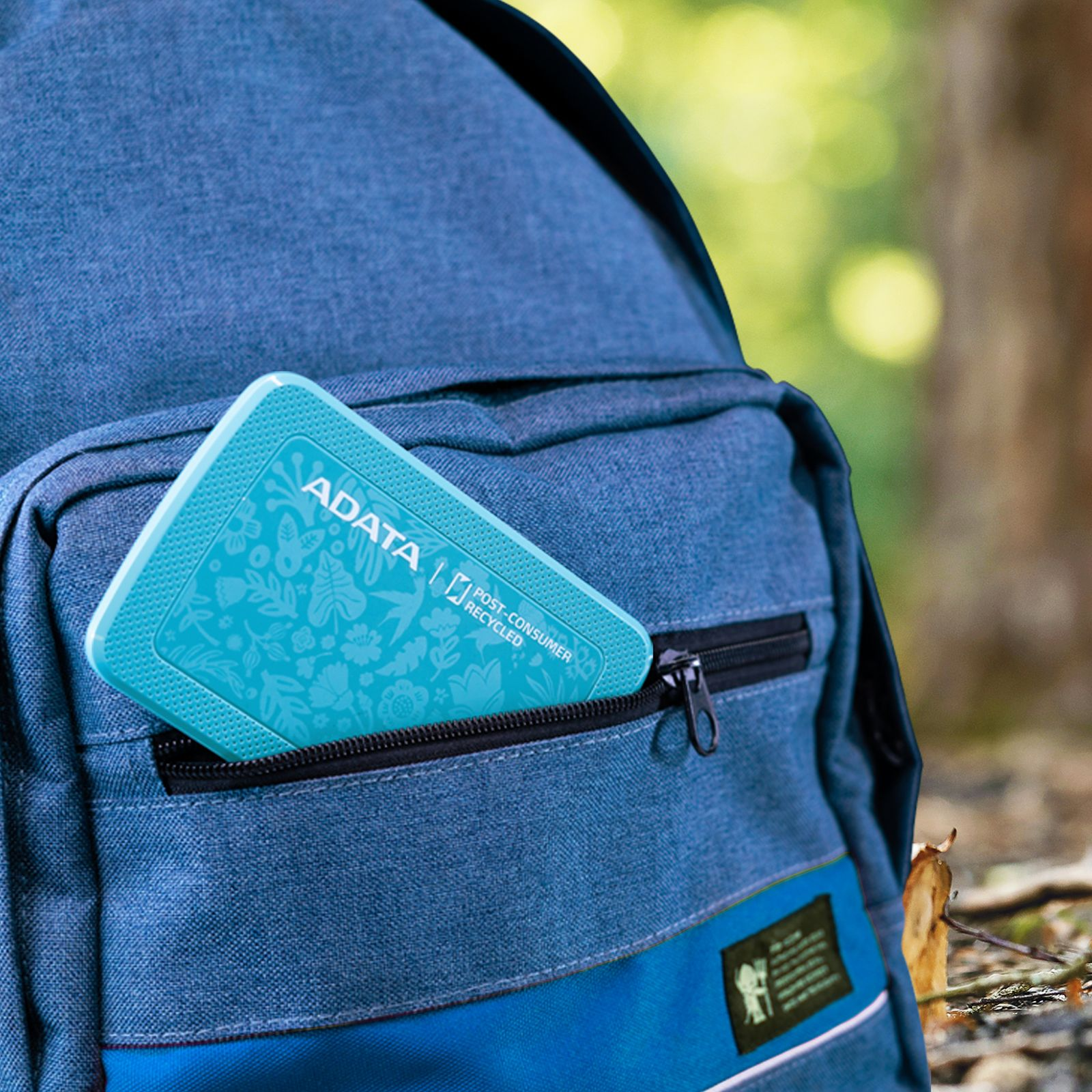 Disco duro externo turquesa en el bolsillo de una mochila azul, en un entorno forestal.