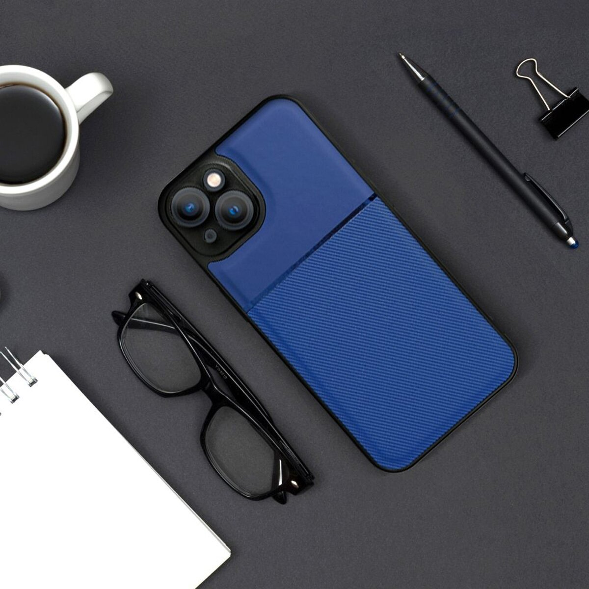 Draufsicht auf eine blaue Handyhülle, Brille, Stift, Notizblock und eine Tasse Kaffee. Alles auf grauer Oberfläche.