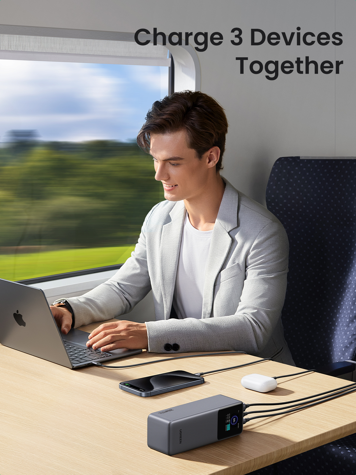 Mężczyzna ładuje laptopa, telefon i białe urządzenie. Jest w pociągu z rozmytym tłem.