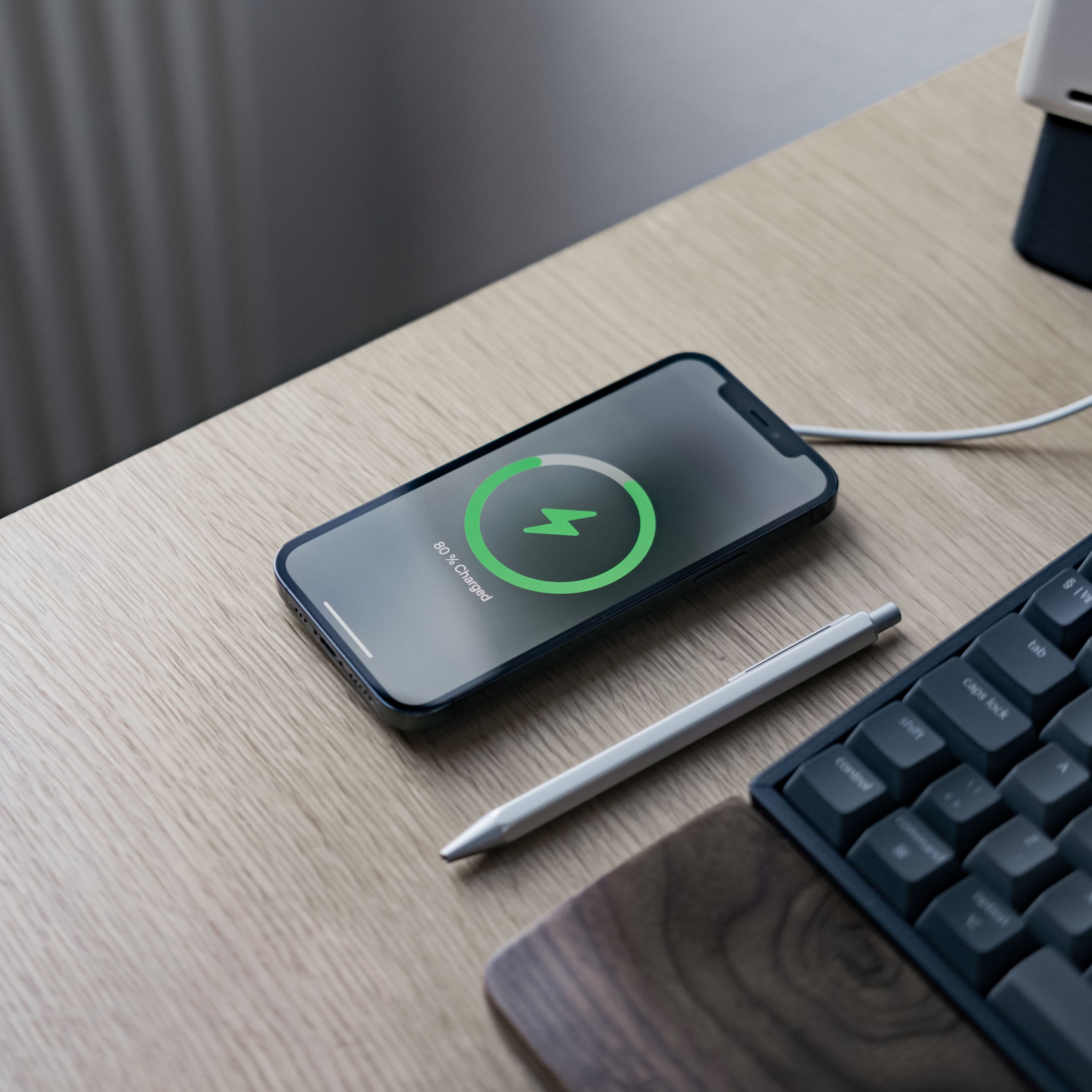 Telefon ładujący się na podkładce bezprzewodowej, 80% naładowania. Ołówek i klawiatura w pobliżu.