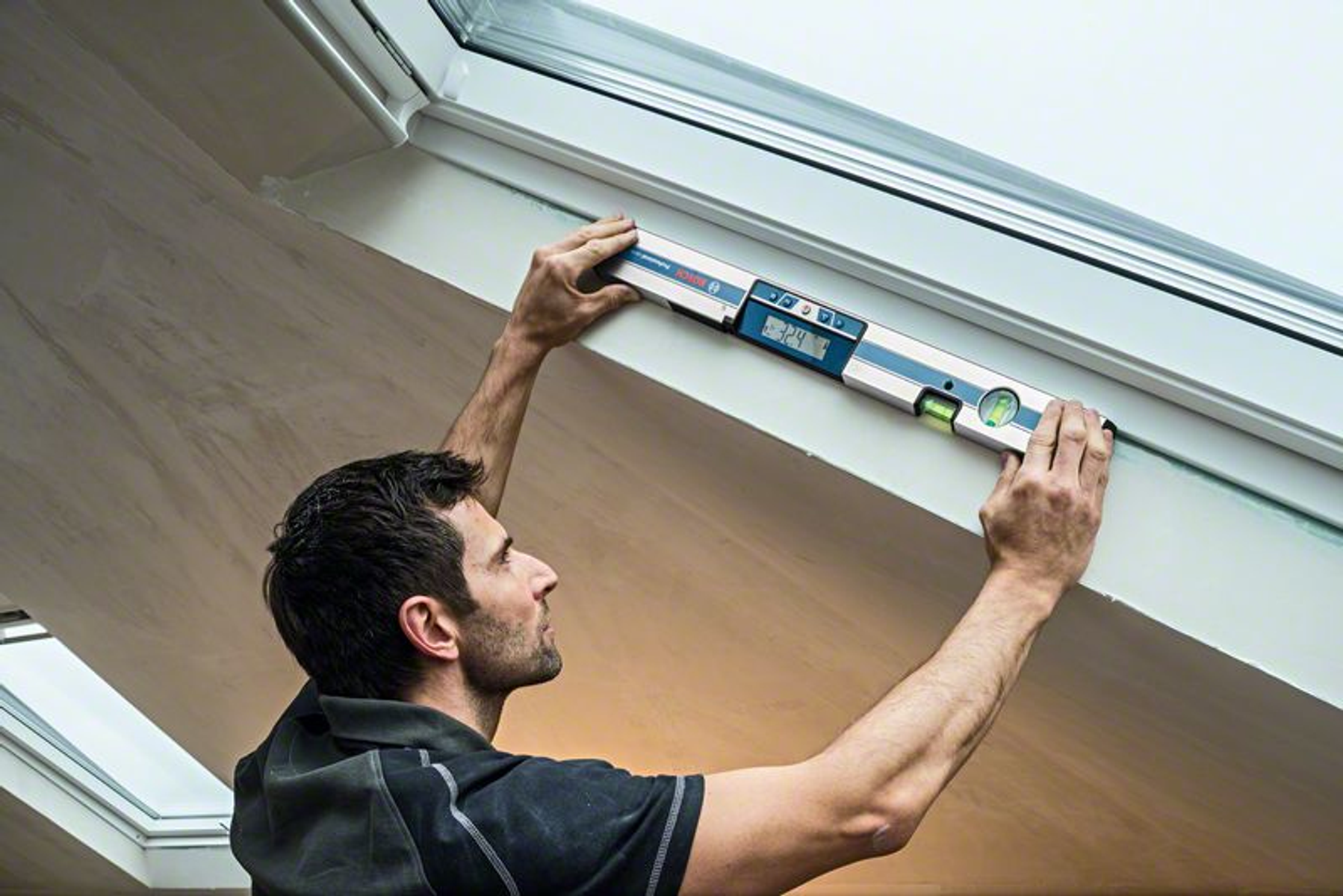 Ein Mann hält eine Wasserwaage an einem weißen Rahmen, wahrscheinlich einem Fensterrahmen, bei Tageslicht.