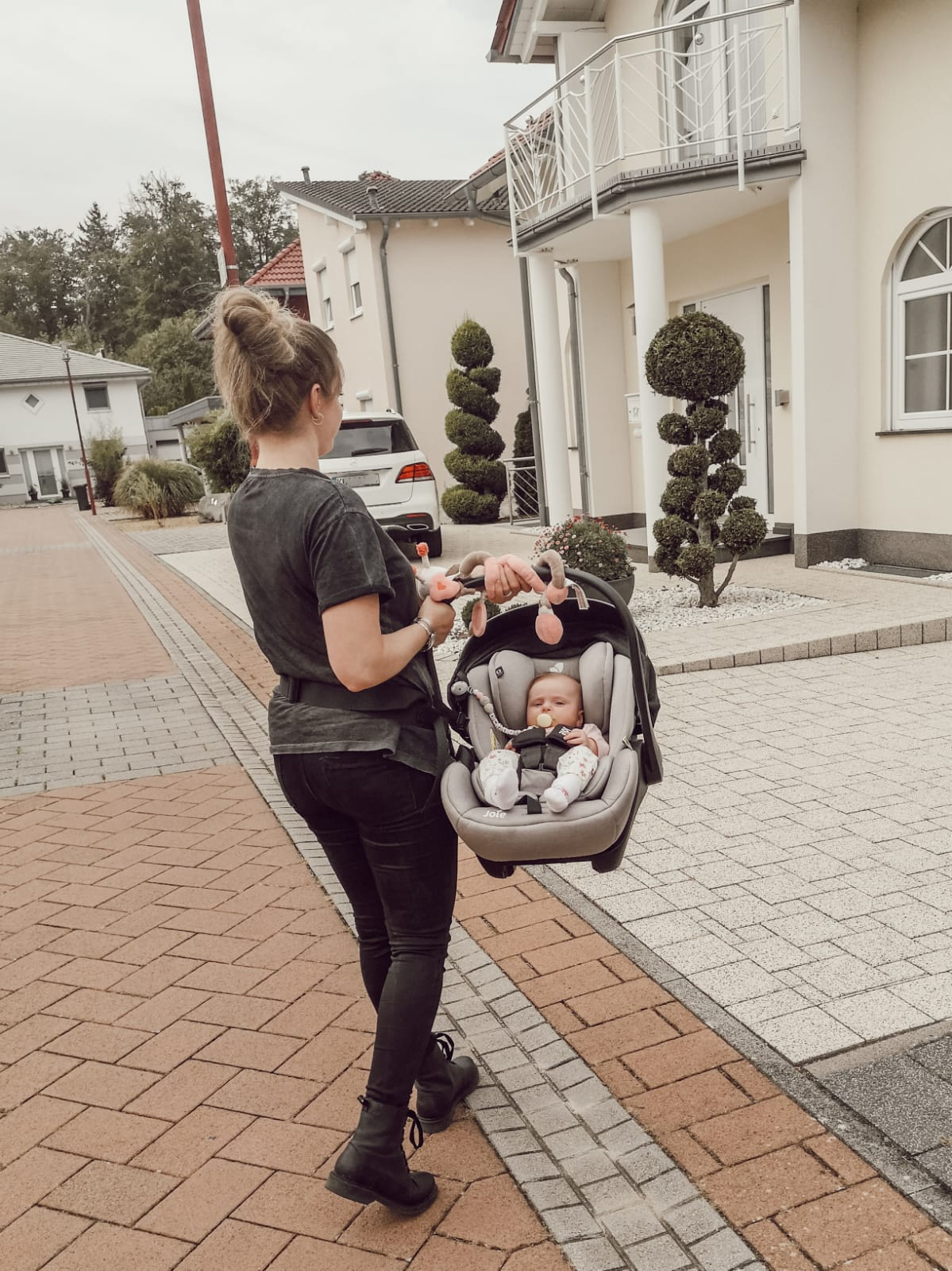 Frau trägt Baby im Autositz auf einer Ziegelstraße, Häuser im Hintergrund.