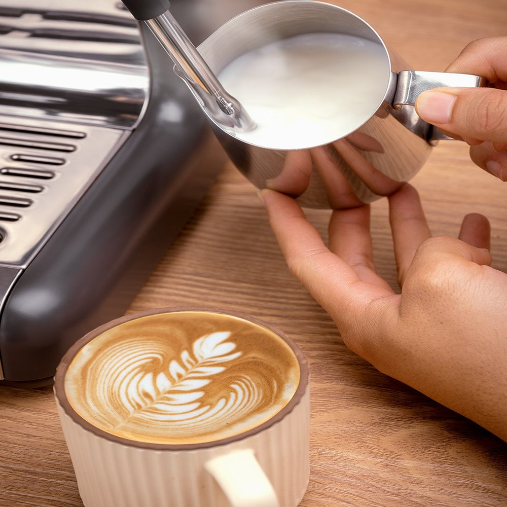 Persona vertiendo leche en una taza de café con arte latte, vista de cerca.