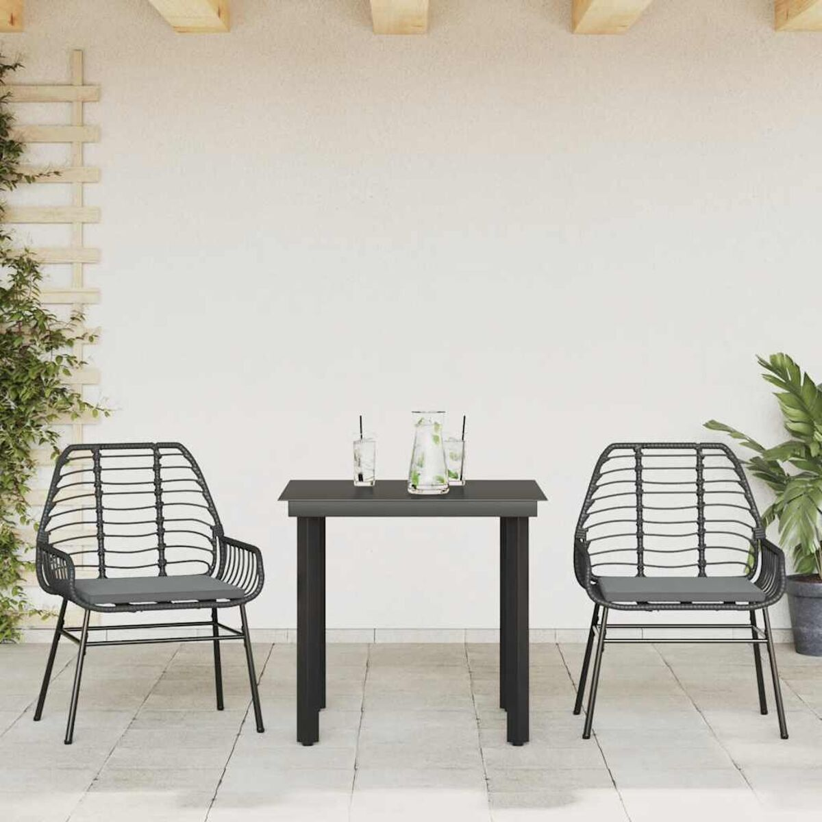 Set da pranzo all'aperto: tavolo nero, due sedie. Parete bianca e piante sullo sfondo.