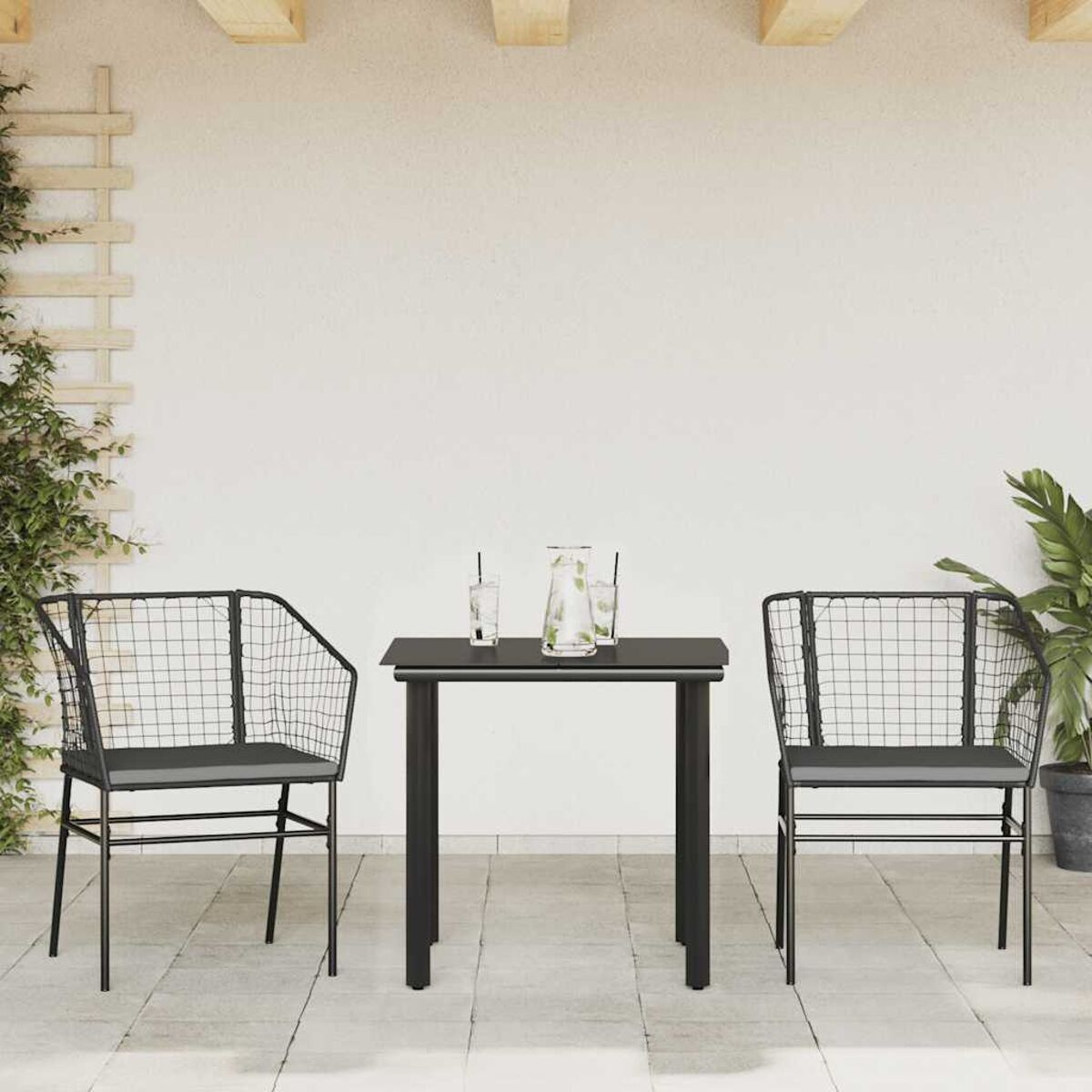 Terrassenset: Tisch, zwei Stühle. Schwarze Metallrahmen, Korbsitze. Hintergrund: weiße Wand.