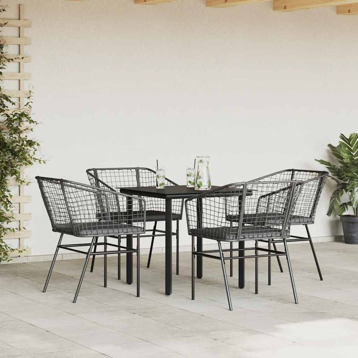 Un tavolo nero quadrato e quattro sedie a rete metallica su un patio, con piante e un muro bianco sullo sfondo.