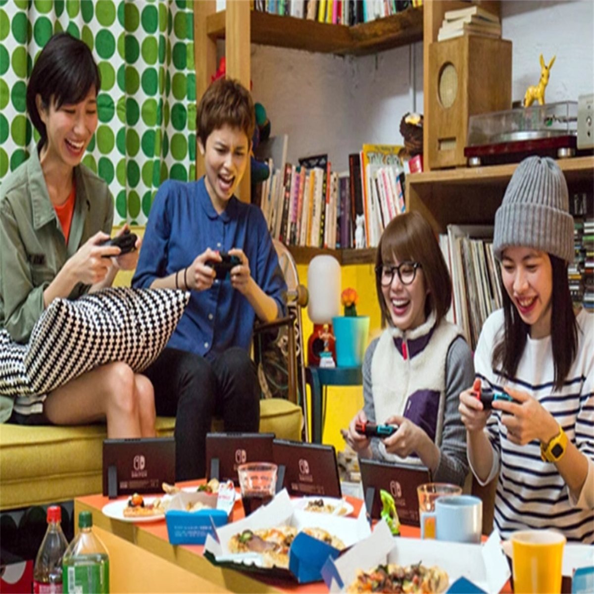 Vier Frauen spielen Videospiele auf einem Sofa, mit Pizza und Snacks auf dem Tisch. Fröhliche Gesichter.
