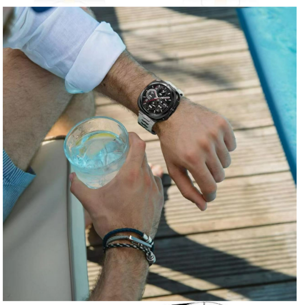 Polso maschile con orologio, che tiene un bicchiere d'acqua. Ponte di legno e piscina sullo sfondo.