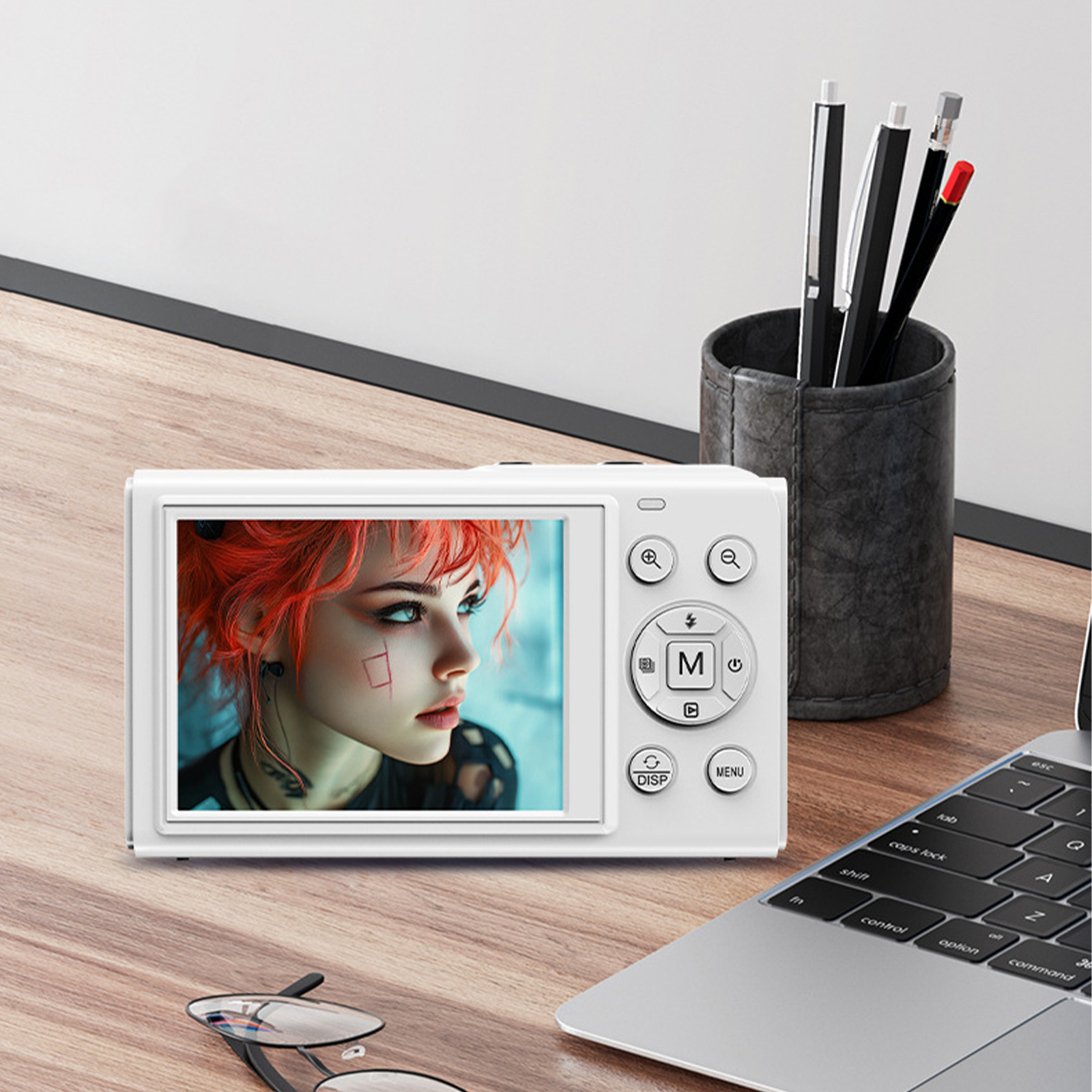 Eine weiße Digitalkamera, die ein Foto eines Frauengesichts anzeigt, mit Stiften und einem Laptop auf einem Holztisch.