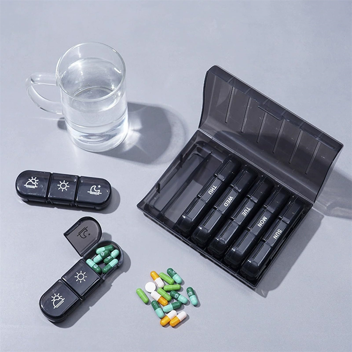 Tabletten-Organizer mit Pillen und einem Glas Wasser auf grauer Oberfläche. Einige Pillen sind draußen.