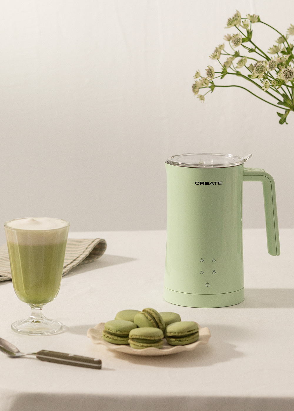 Un espumador de leche verde, un vaso de matcha latte y macarons en mesa blanca.