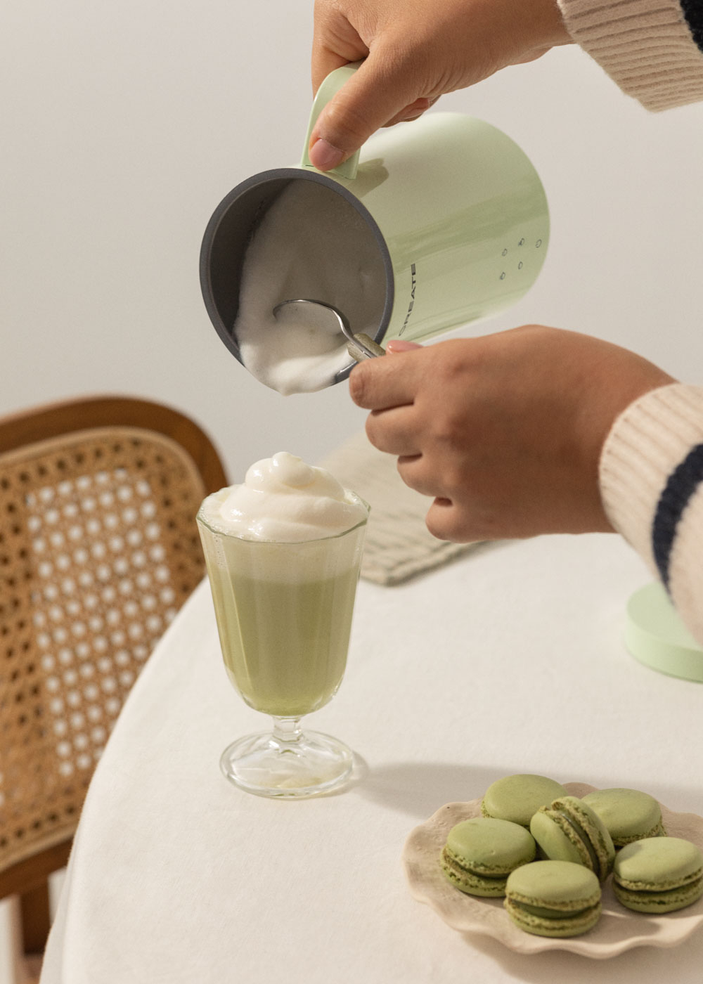 Manos vertiendo bebida espumosa en un vaso. Al lado, macarrones verdes.