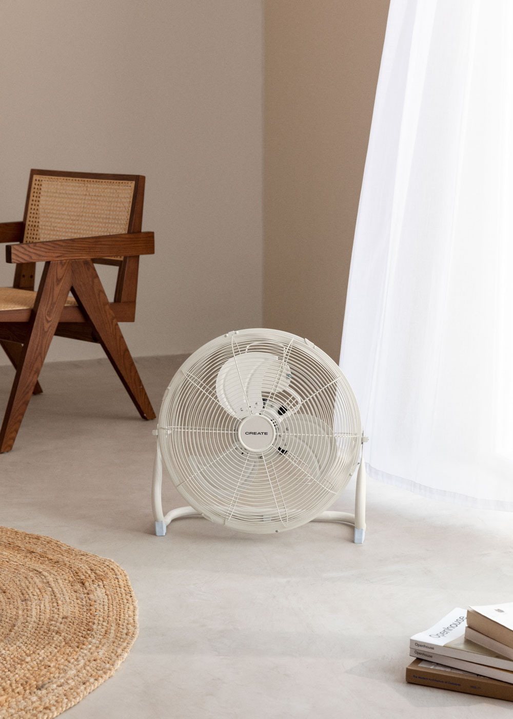 Ventilador blanco en el suelo, frente a una ventana. Una silla de madera y una alfombra están en el fondo.