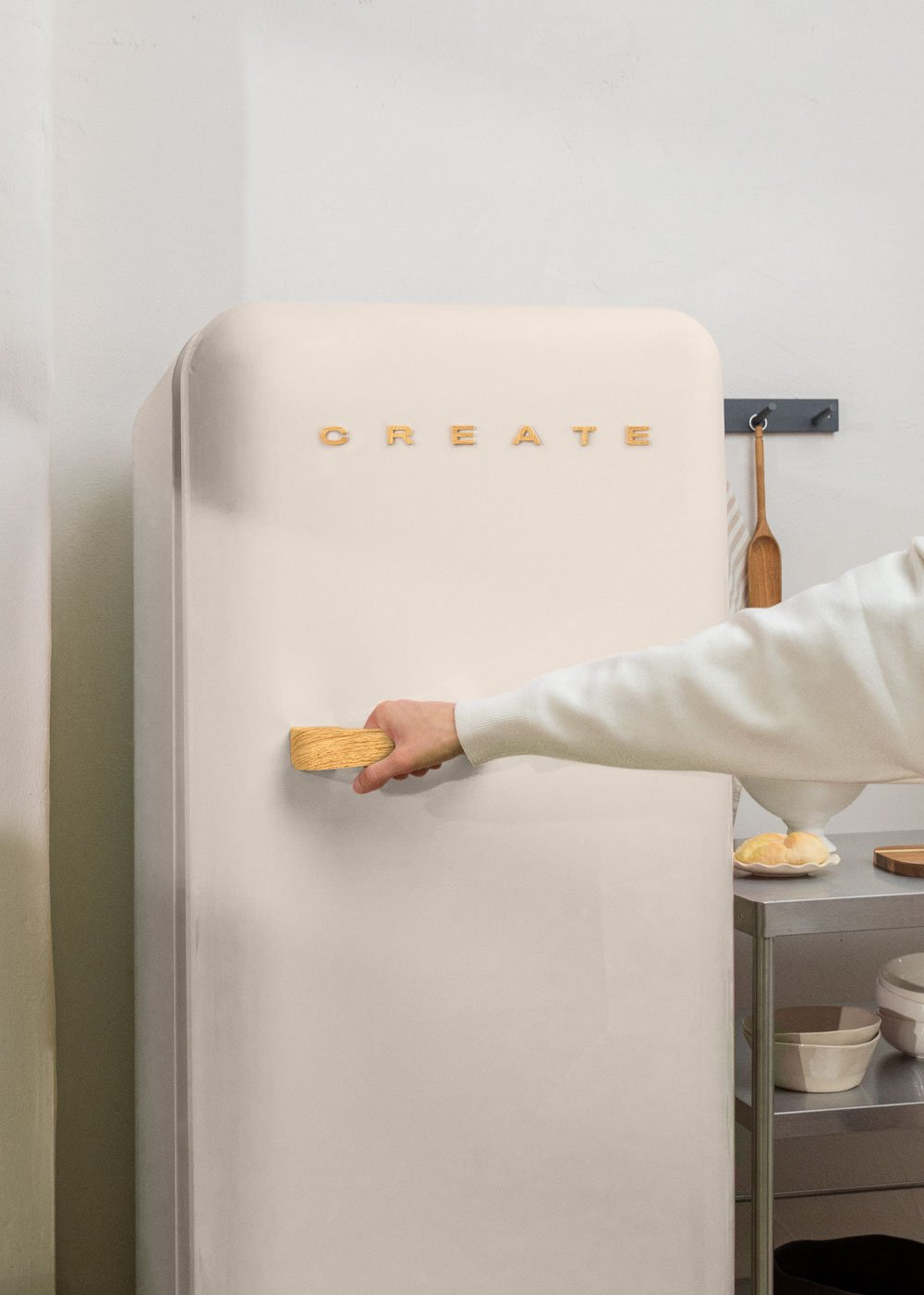 Una persona abre una nevera color crema con la palabra 'CREATE'. El tirador es de madera.