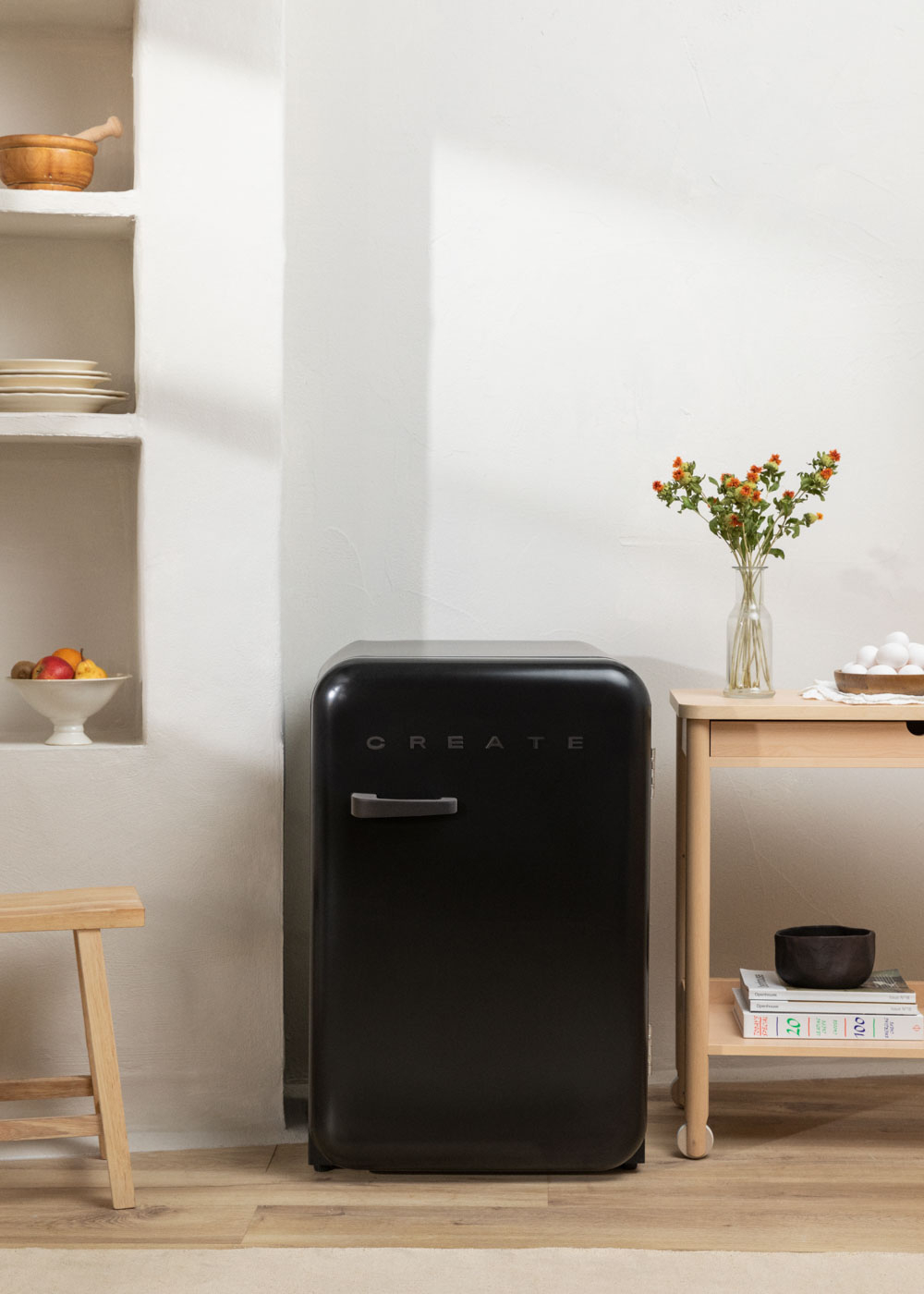 Mini refrigerador negro con letras 'CREATE', en una cocina con madera, flores y estantes.