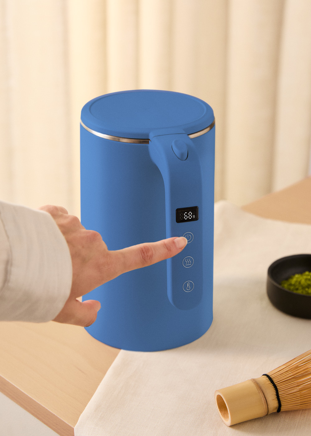 Una mano señala el botón de una tetera eléctrica azul. Al lado hay un cuenco de matcha y un batidor de bambú.