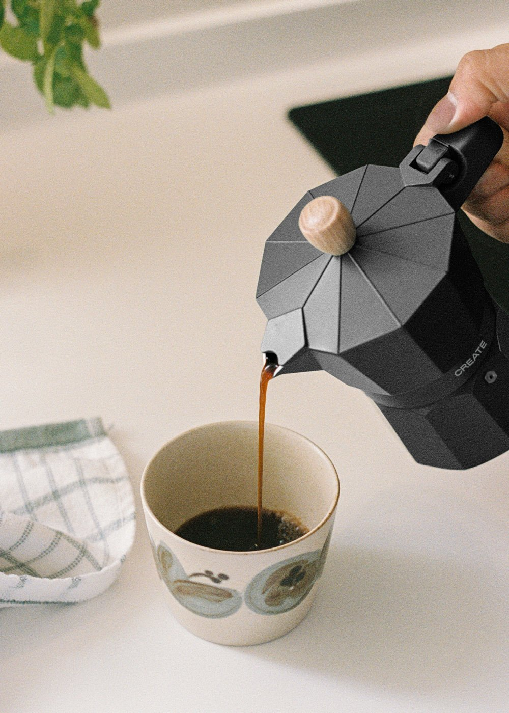 Café vertiéndose de una cafetera moka negra en una taza. Encimera blanca y un paño en el fondo.