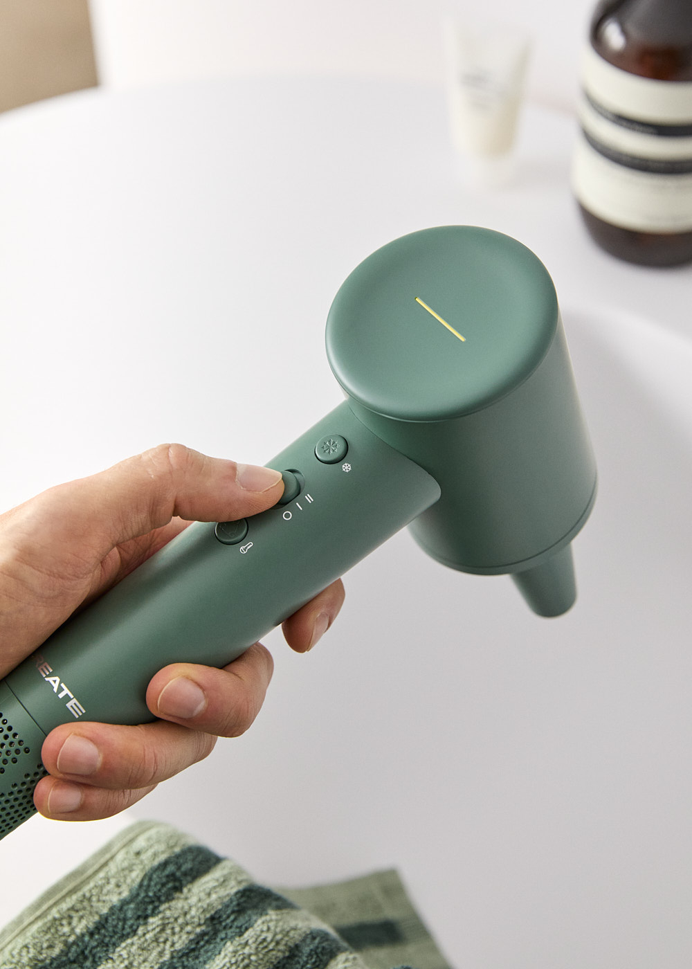 Se sostiene un secador de pelo verde, un dedo pulsando un botón. Fondo blanco.