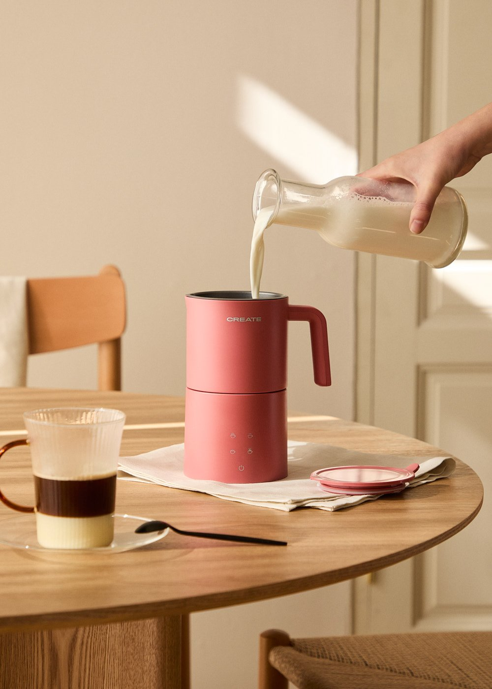 Persona vertiendo leche en un aparato CREATE rosa en mesa de madera. Café cerca.