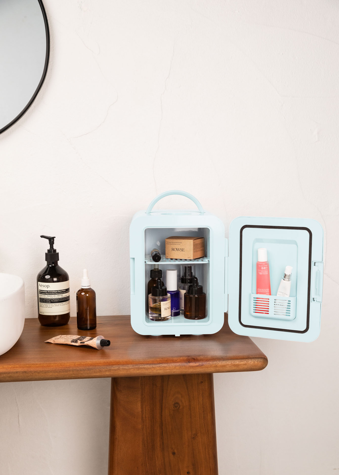 Mini nevera azul abierta, con productos de belleza dentro. En una mesa de madera. Fondo de baño.