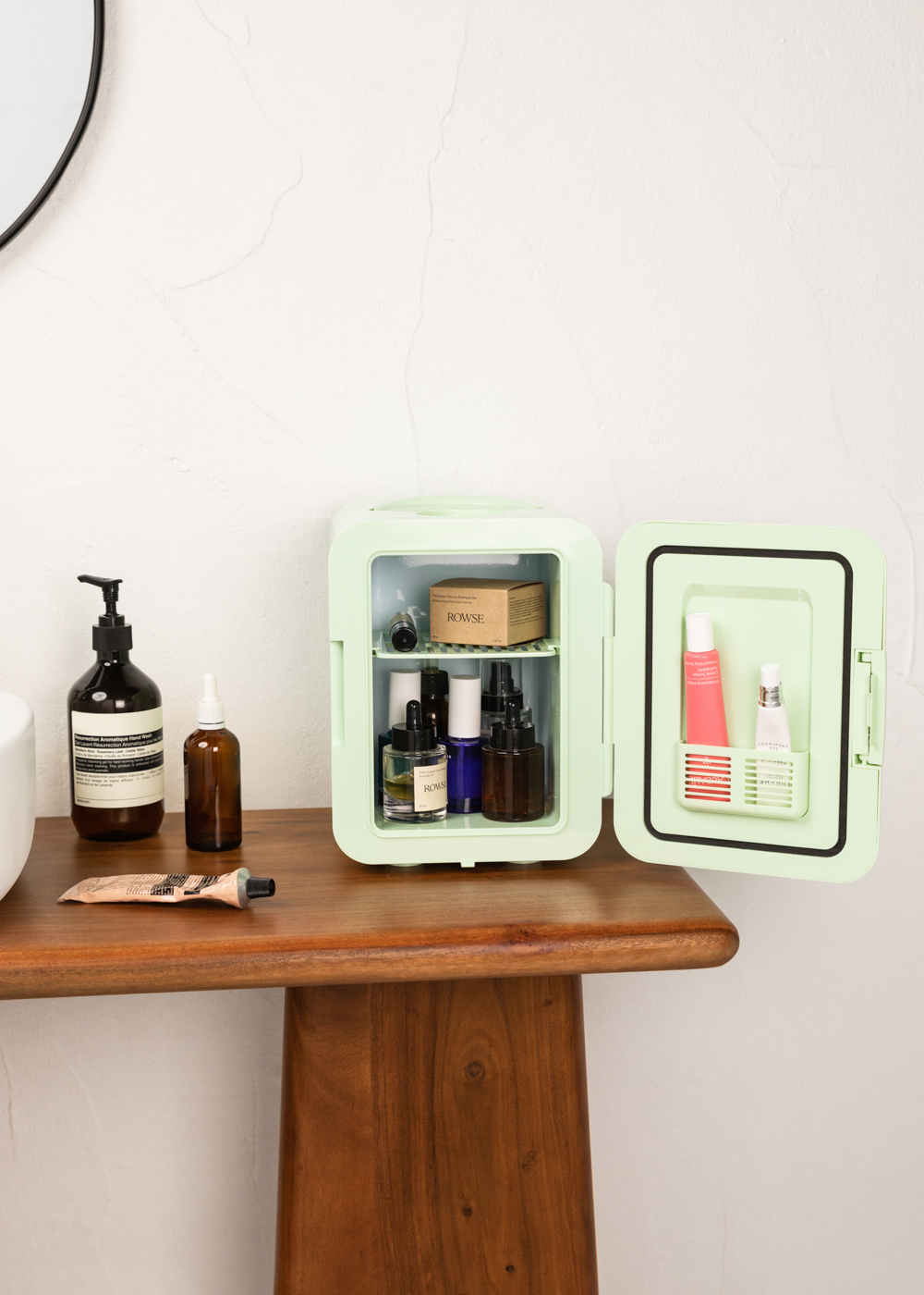 Mini nevera abierta, con productos de belleza dentro. En una mesa de madera con productos y espejo cerca.