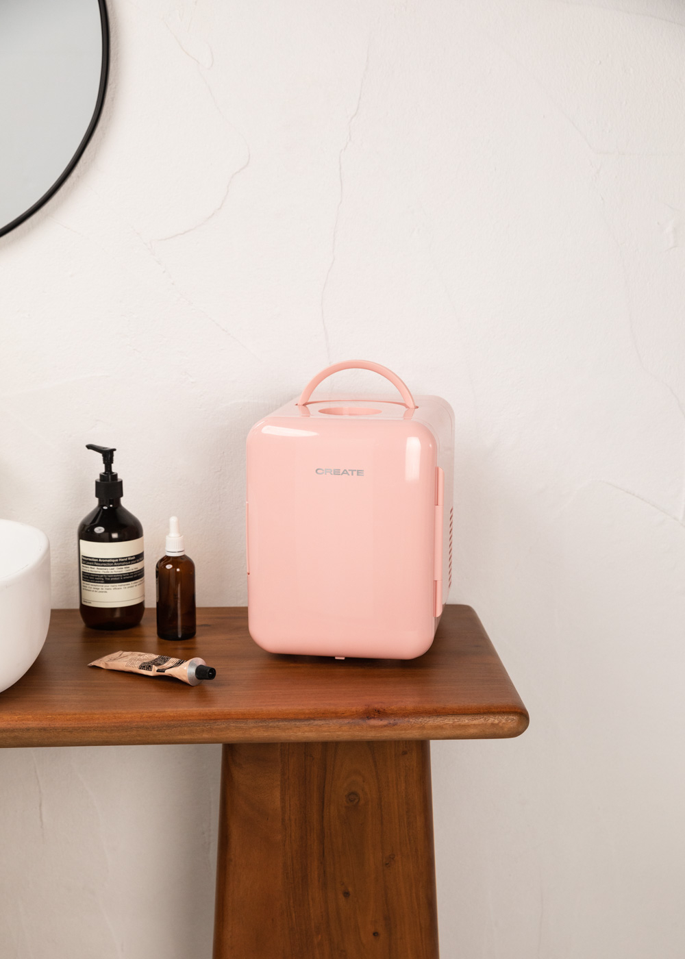 Una mini-nevera rosa sobre una mesa de madera con productos para el cuidado de la piel y un lavabo blanco al fondo.