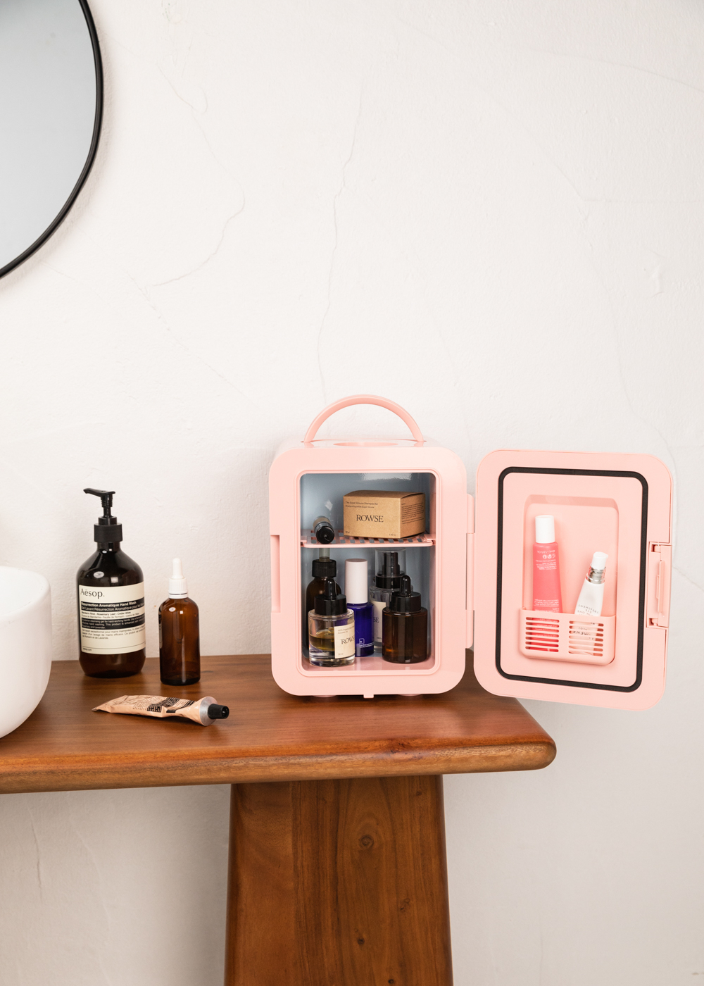 Un mini-frigorífico rosa con la puerta abierta que revela productos para el cuidado de la piel en una mesa de madera junto a un lavabo.