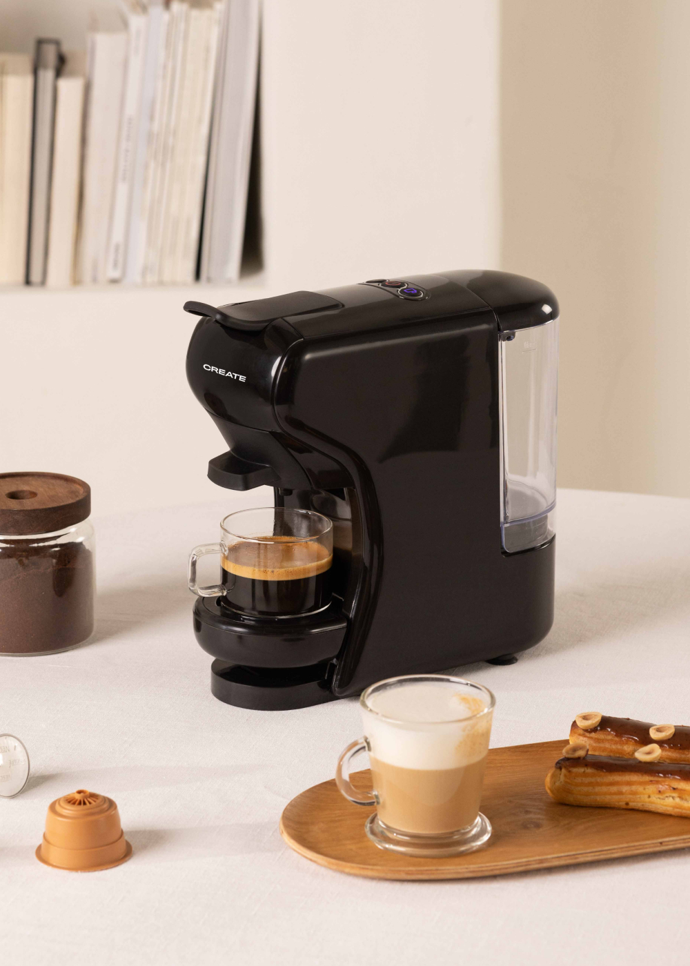 Cafetera negra con café en vaso. Cápsulas, café molido y pasteles en mesa.